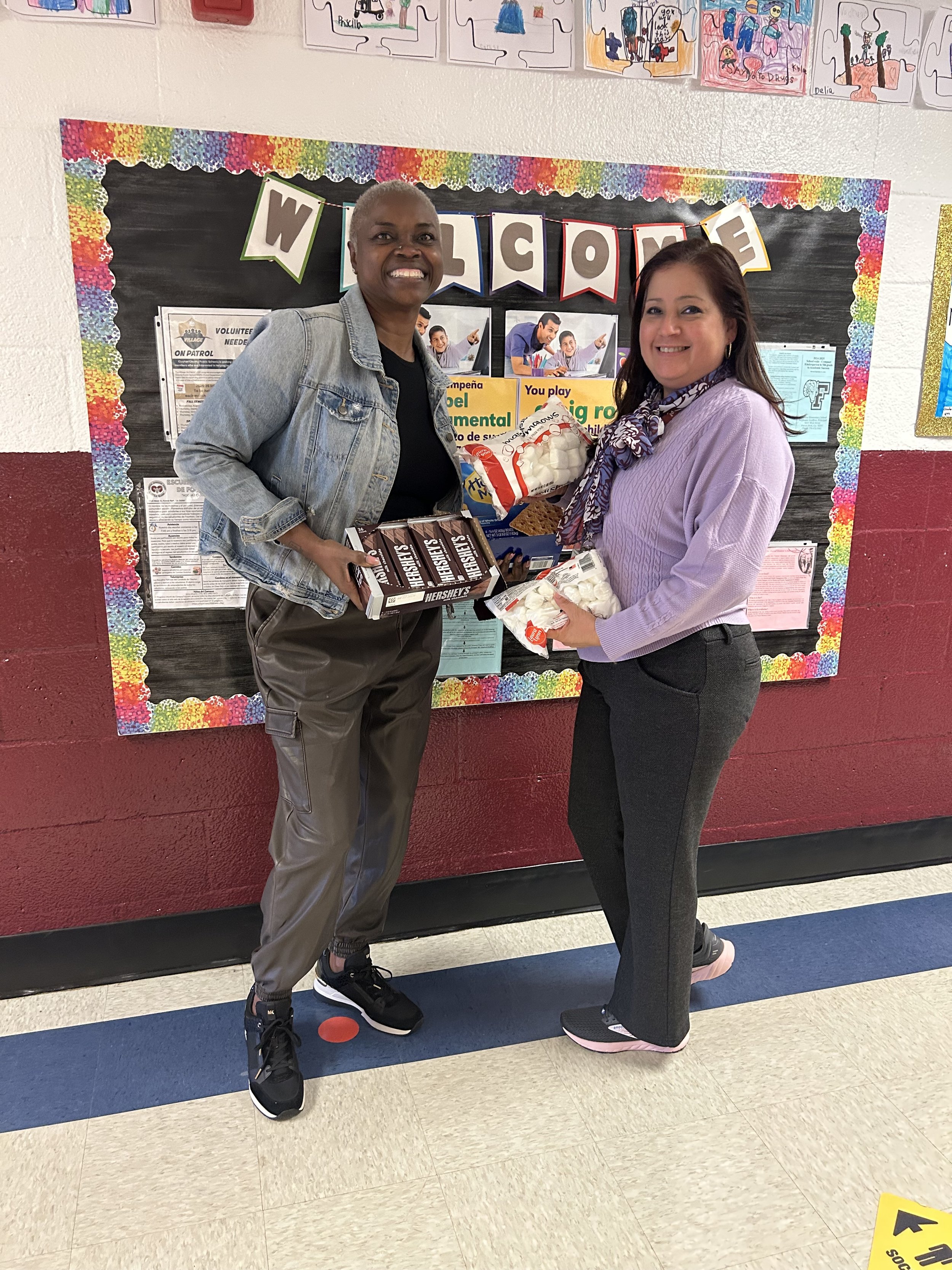 Two women standing in a school hallway, smiling and holding snacks, in front of a colorful bulletin board that has a banner saying "WELCOME." The woman on the left has short gray hair and is wearing a denim jacket, black shirt, gray pants, and sneakers. The woman on the right has brown hair and is wearing a lavender sweater, scarf, gray pants, and sneakers.