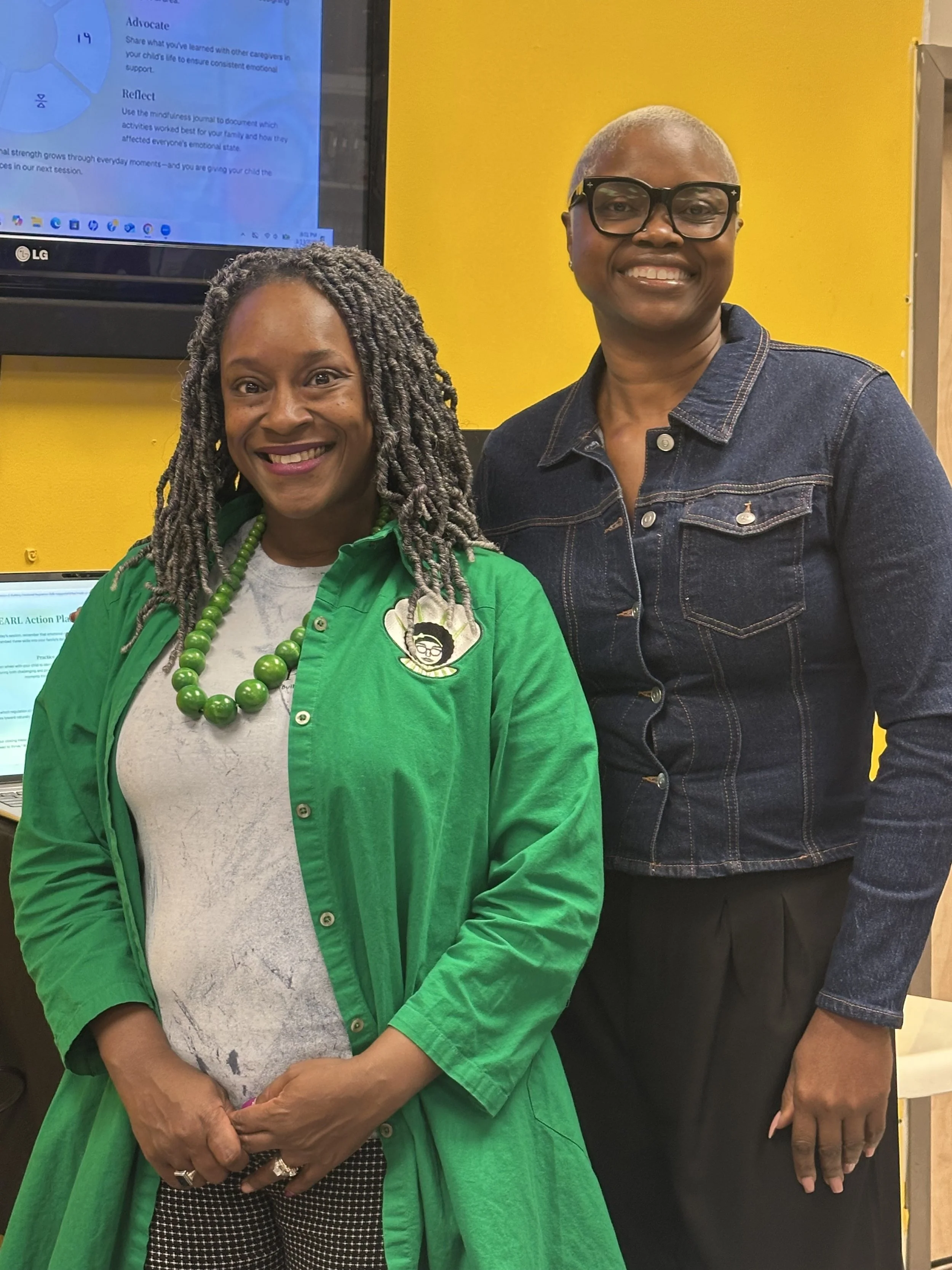 Two women standing side by side, smiling at the camera, against a yellow wall with a monitor in the background.