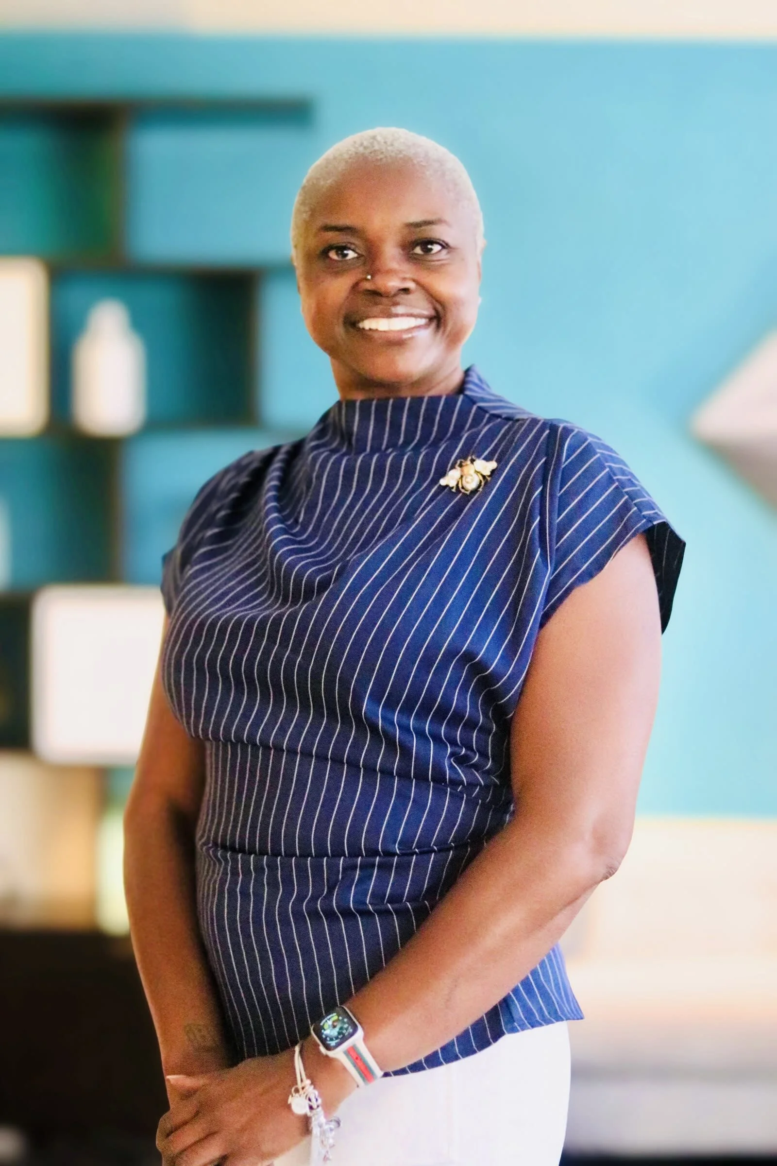 A woman with short blonde hair, wearing a navy blue striped top with a pin, and white bottoms, smiling indoors with a blue wall and shelves in the background.