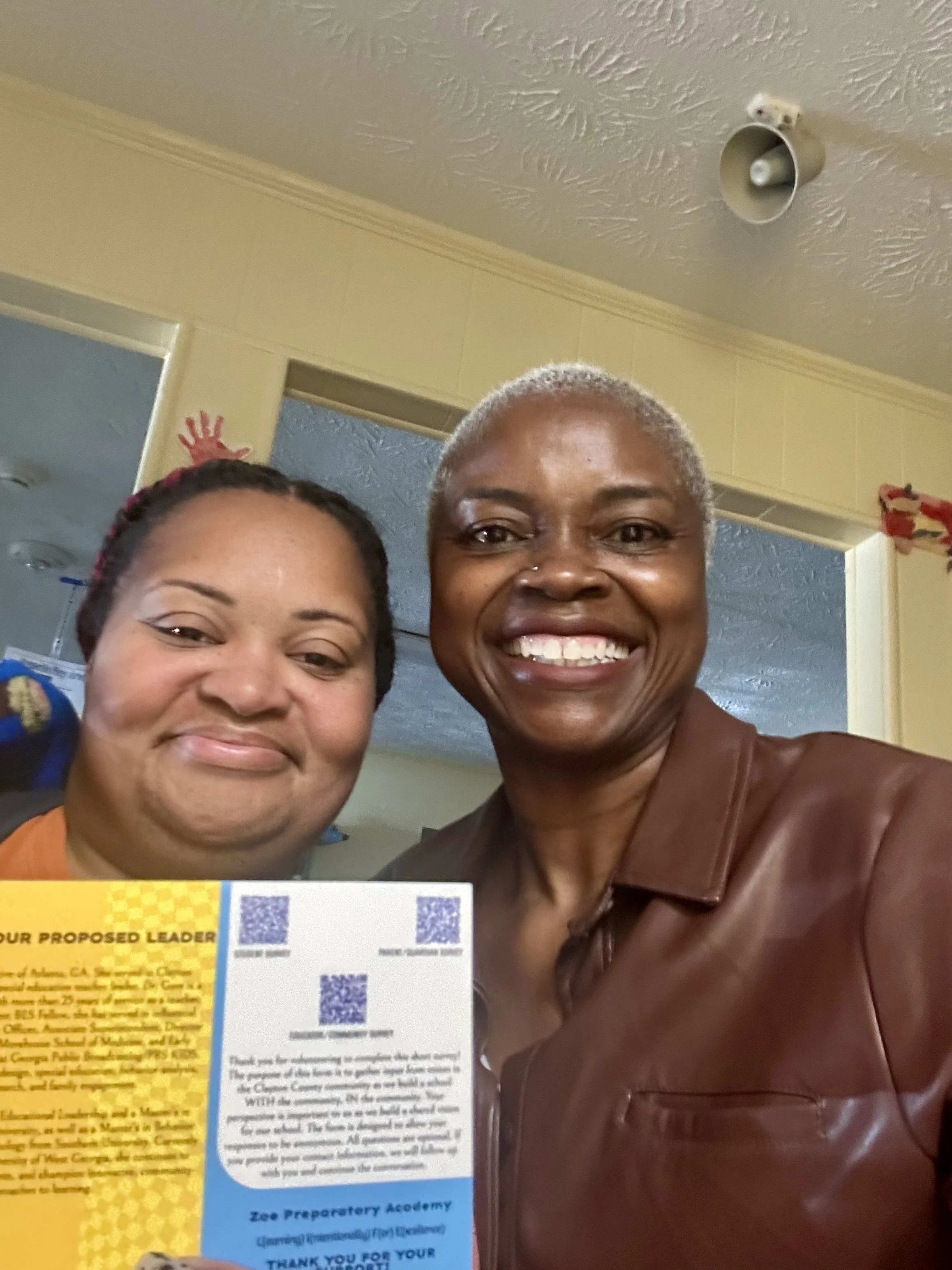 Two women smiling in a selfie, one with short gray hair and the other with dark hair, indoors with a yellow wall and doorway in the background. There are promotional materials and a QR code in the foreground.