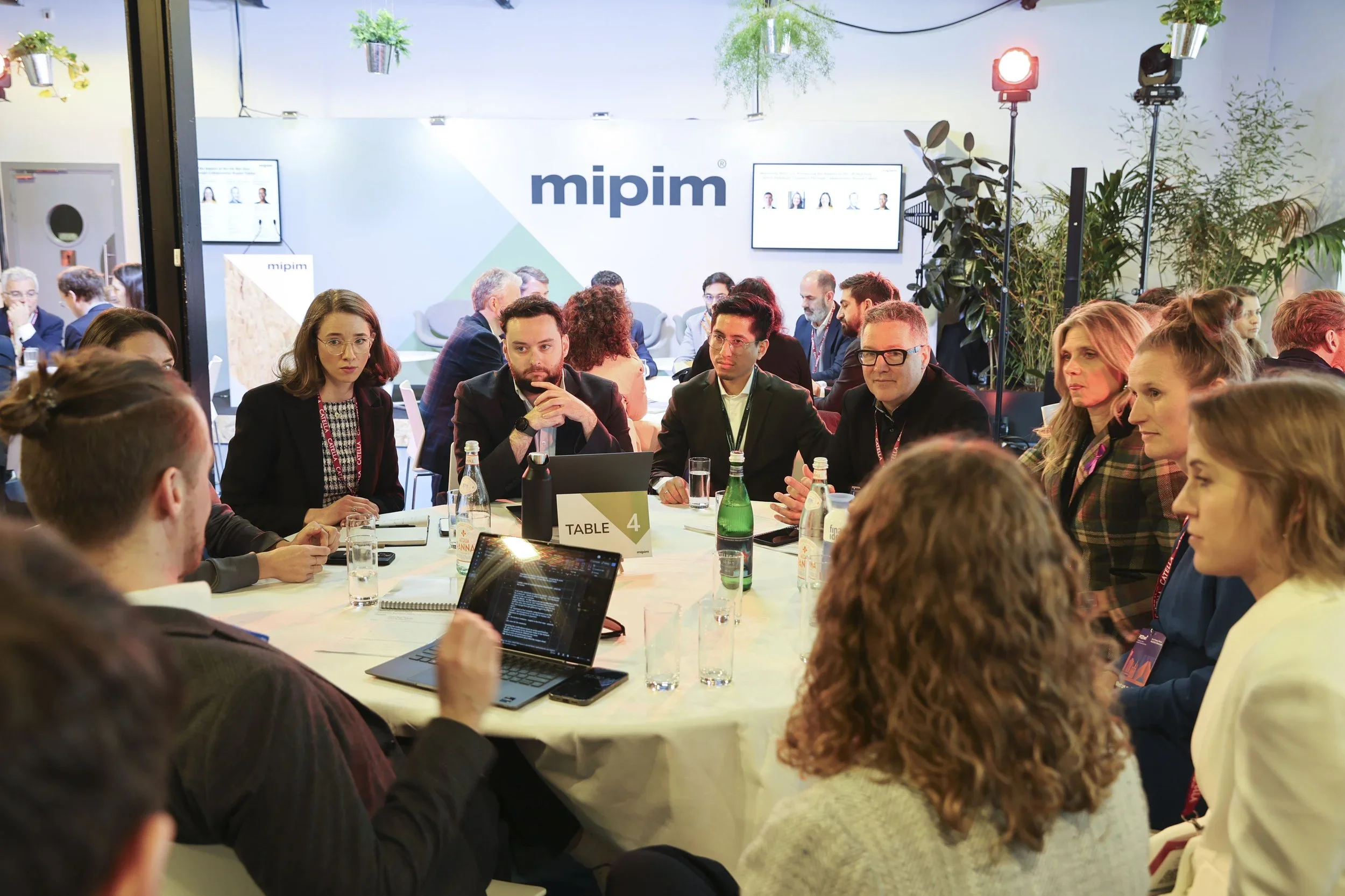 Business professionals seated around a circular table engaged in a discussion at a conference or workshop, with the room branded with 'mipim' signage and large screens in the background