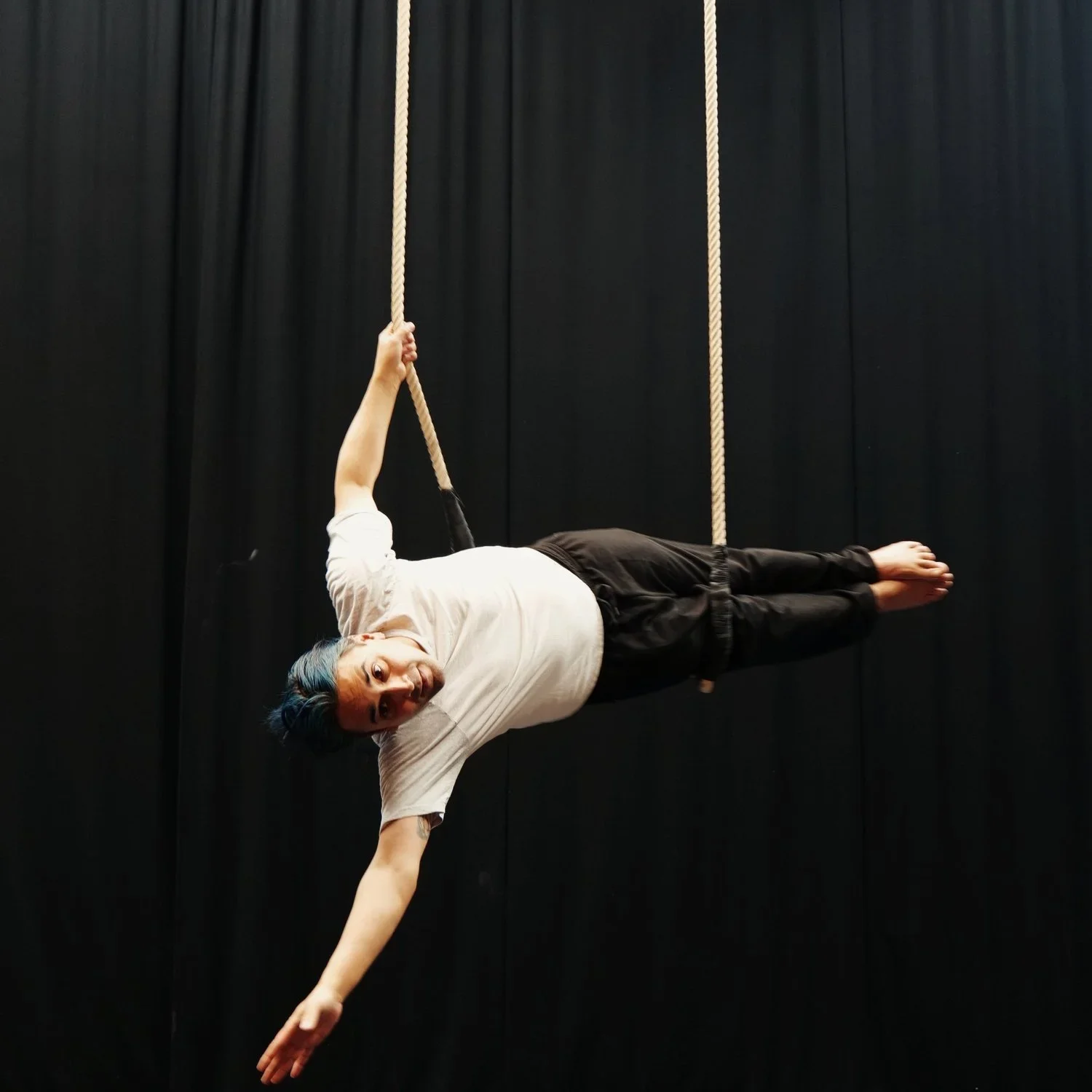 A person hanging horizontally from a climbing rope against a black curtain background, with one arm holding the rope and the body extended sideways.