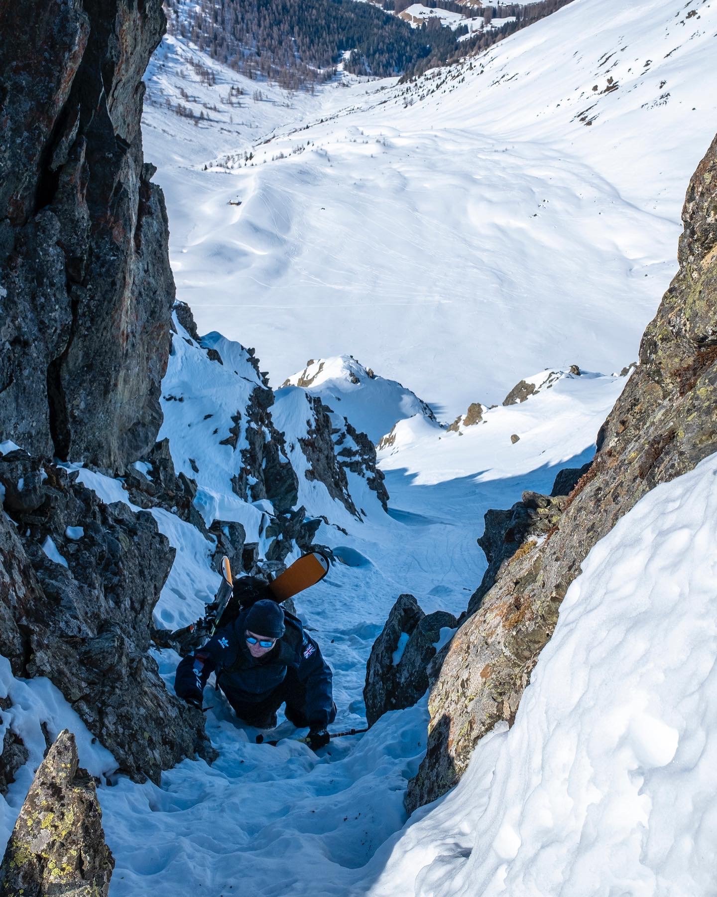A person wearing a blue jacket and sunglasses climbing up a snow-covered, narrow rock crevice in a mountainous area, with snow and trees visible in the background.