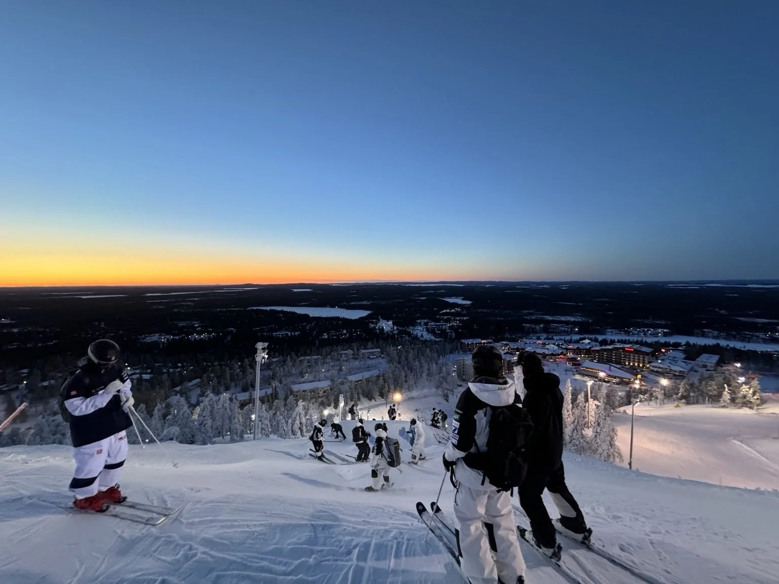 Skiers on a snowy slope during sunset with a view of a town and a forested landscape in the distance, and a colorful sky at dusk.