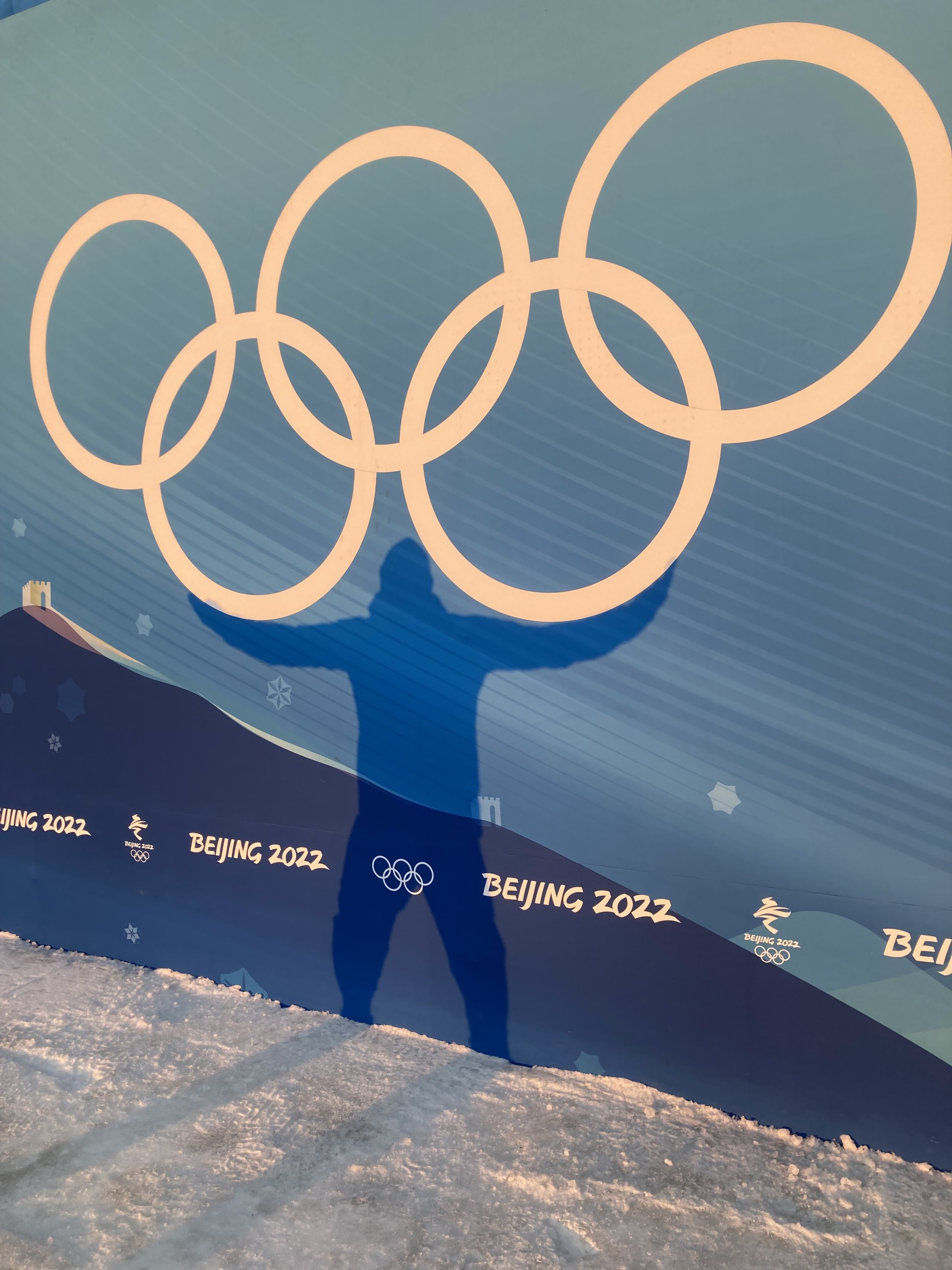 Shadow of a person taking a photo of a large blue wall with the Olympic rings and 'Beijing 2022' written repeatedly on it. The scene is snowy and appears to be outdoors.