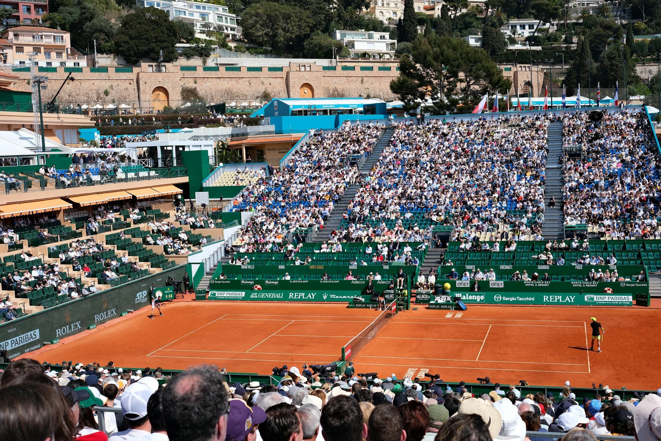 A crowd of spectators seated in green and beige stadium seats watches a tennis match on a clay court surrounded by advertising boards.