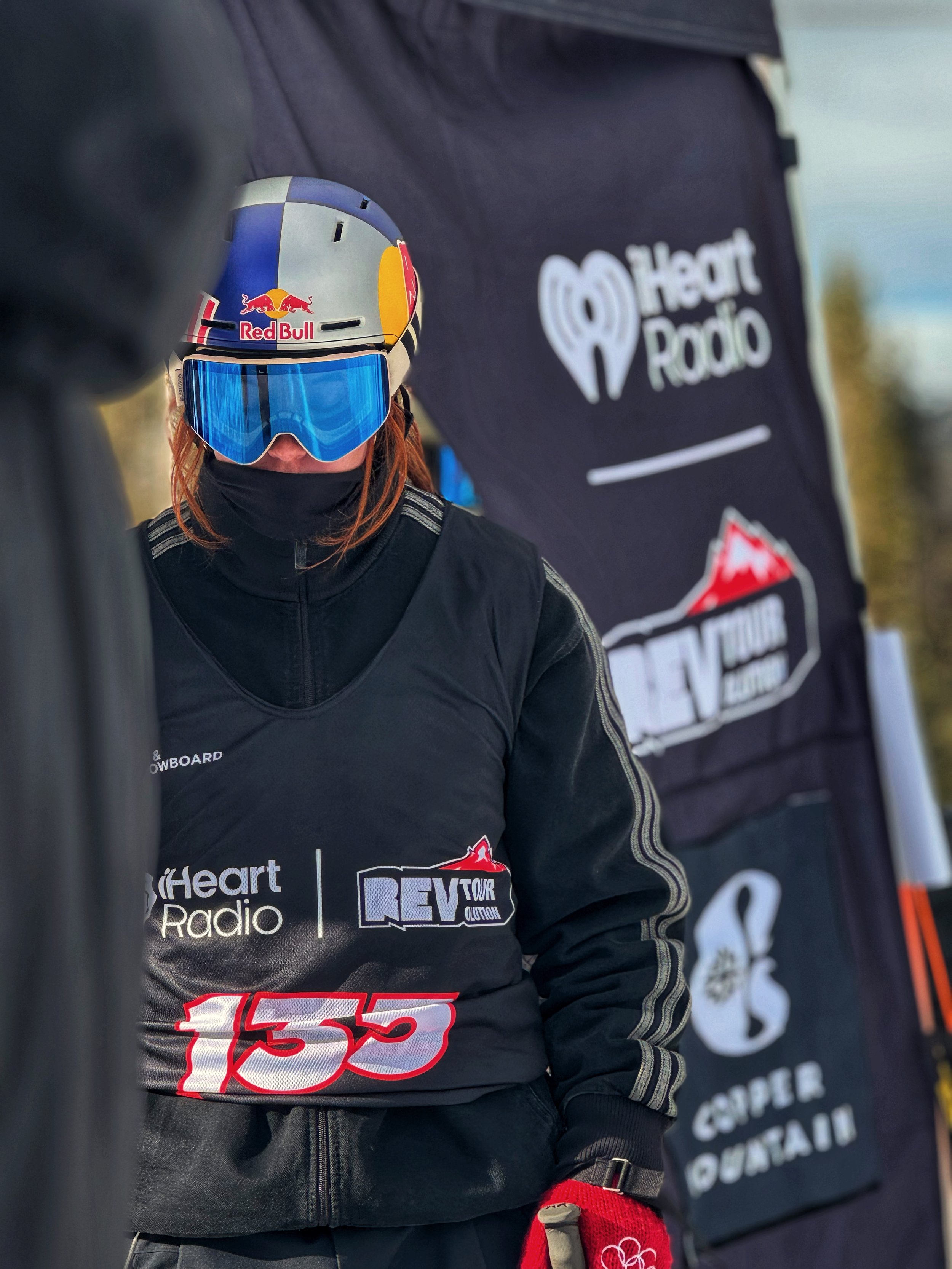 A skier wearing a helmet with Red Bull logo, reflective goggles, and a black ski jacket with logos including iHeart Radio, REV TOUR, and the number 155, standing in front of a black banner with similar logos and a seahorse graphic.