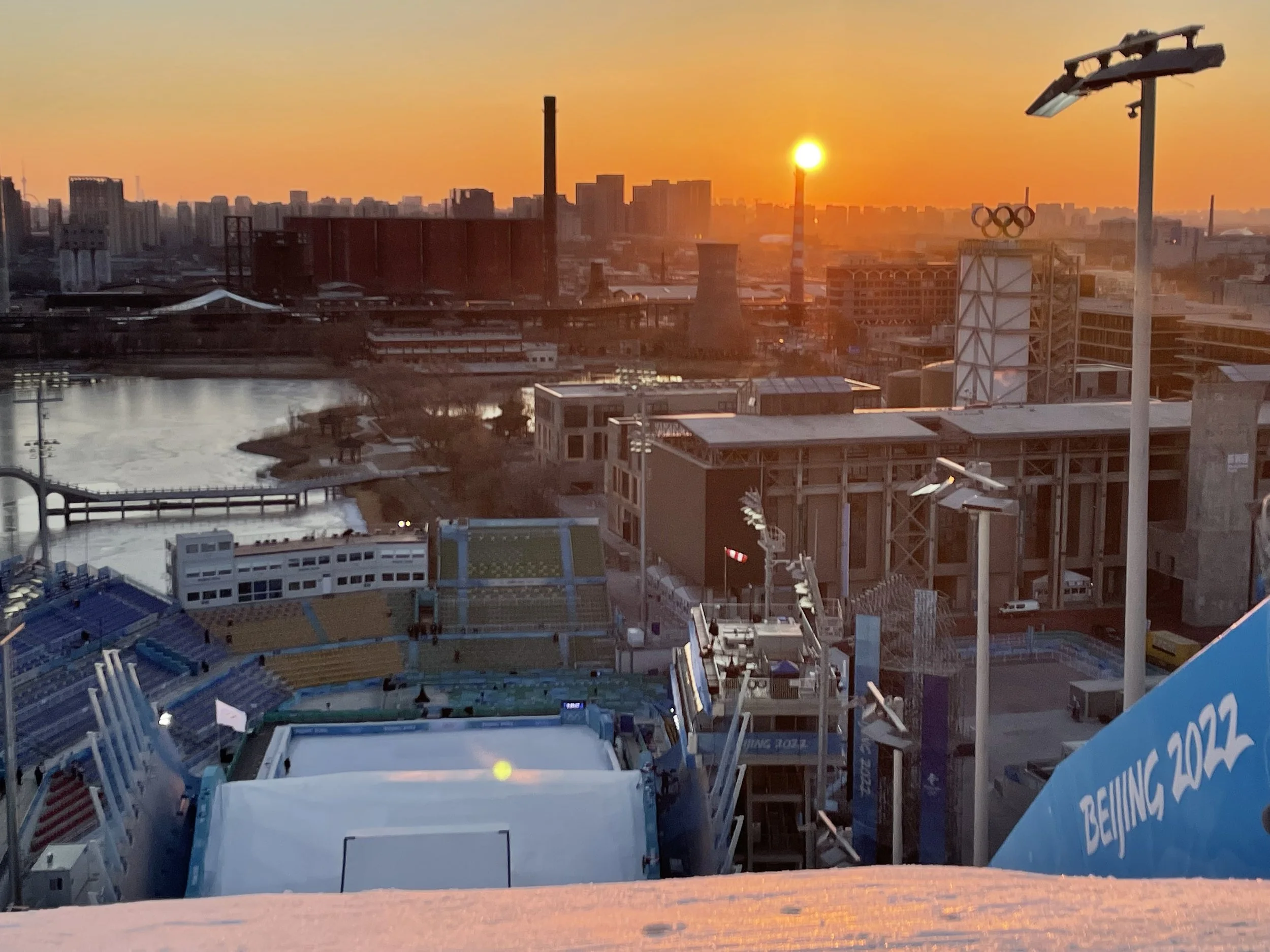 Sunset over a city skyline with tall buildings and a body of water in the foreground. There are sports stadium seating and flags, with the Olympic rings logo visible near the right side of the image.
