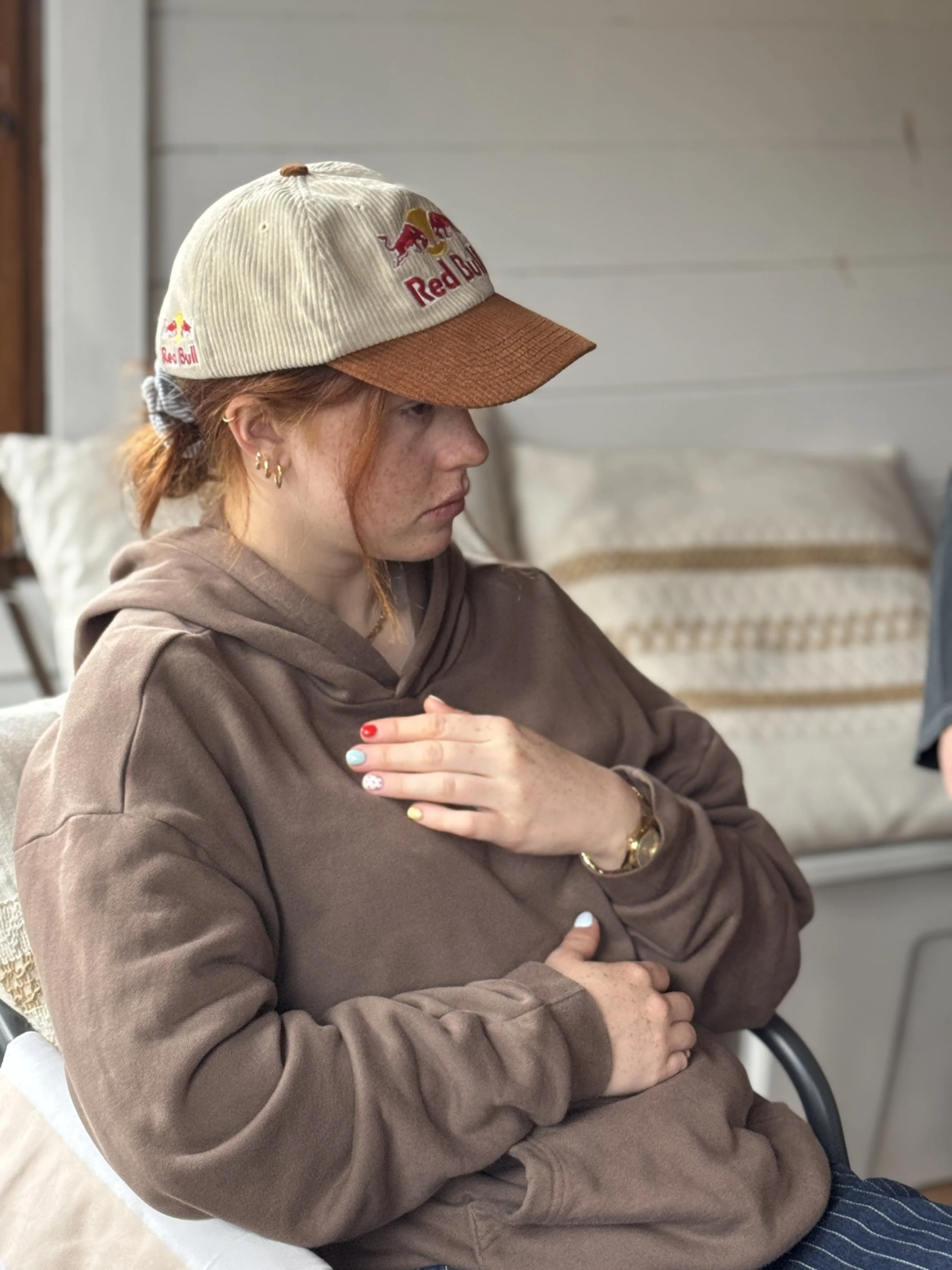 A woman with red hair wearing a beige and brown Red Bull cap, gold earrings, a gold bracelet, and a brown hoodie sitting on a chair with her arms crossed. She has painted nails and is in a room with white walls and a bed with patterned bedding in the background.