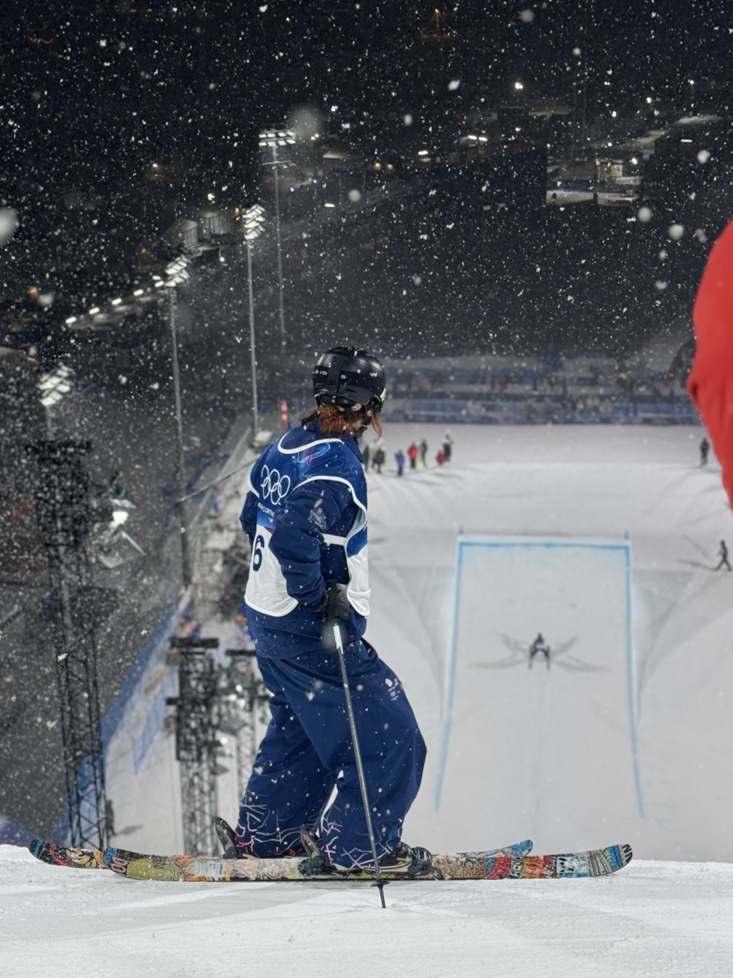 Olympic skier standing on snow, wearing a blue and white outfit and black helmet, preparing for a competition at night with snow falling.