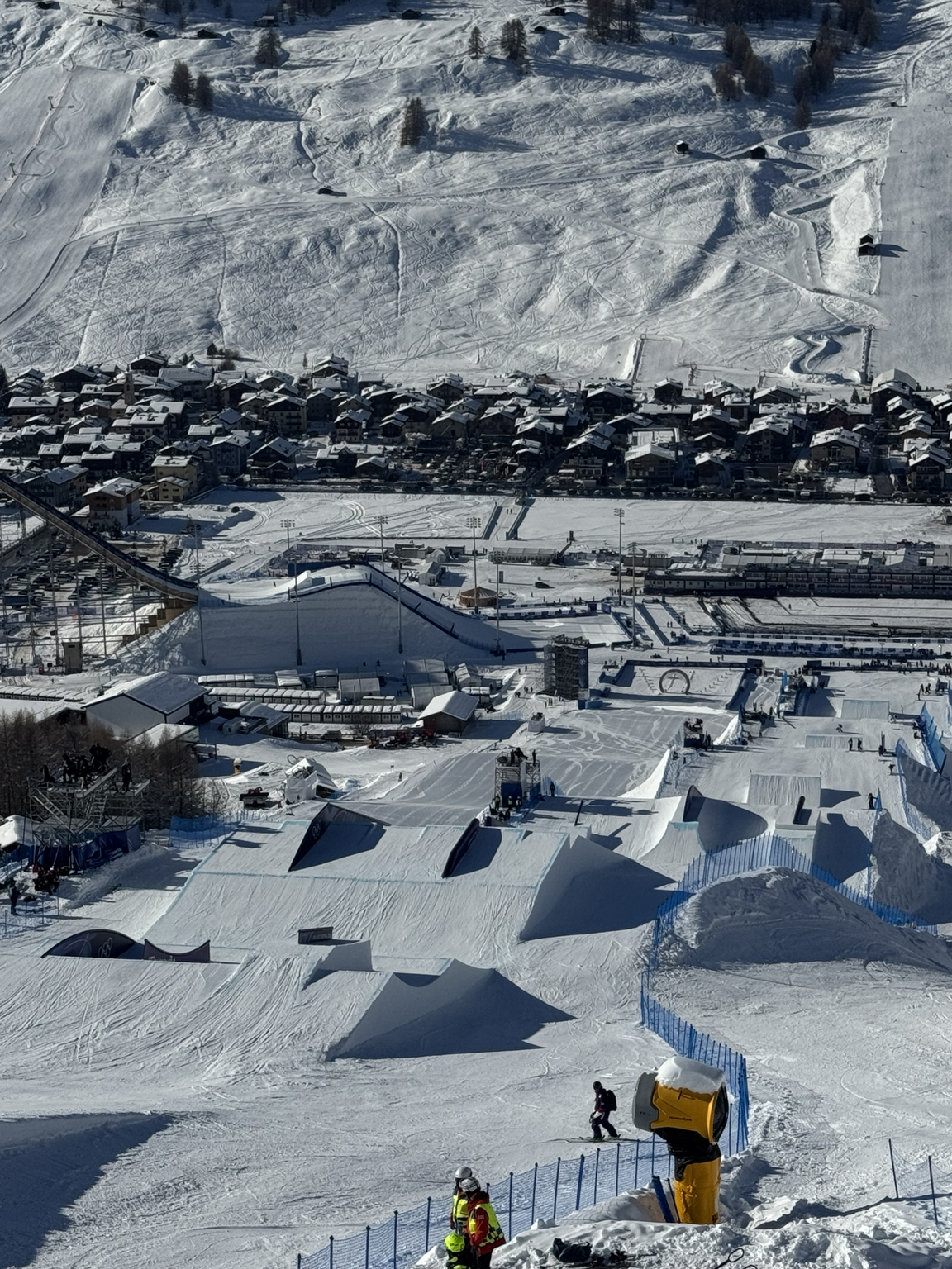 A snowy winter landscape at a ski resort with ski jumps, ski slopes, and skiers, bordered by a town with houses and a mountain with ski trails in the background.