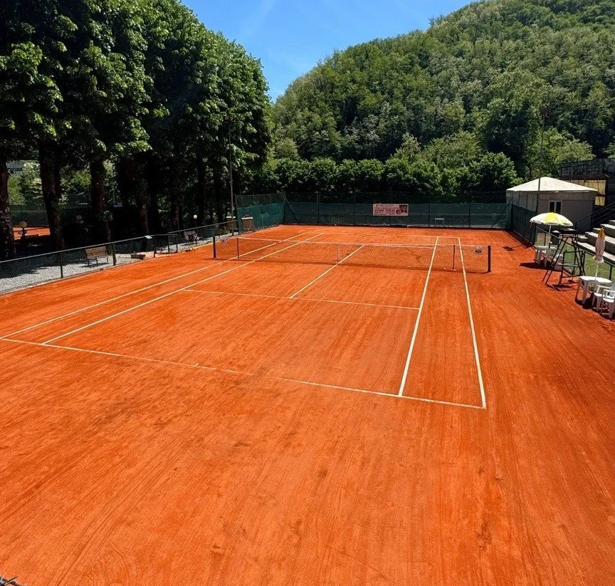 An outdoor clay tennis court surrounded by trees and greenery, with a blue sky and some shade structures on the side.