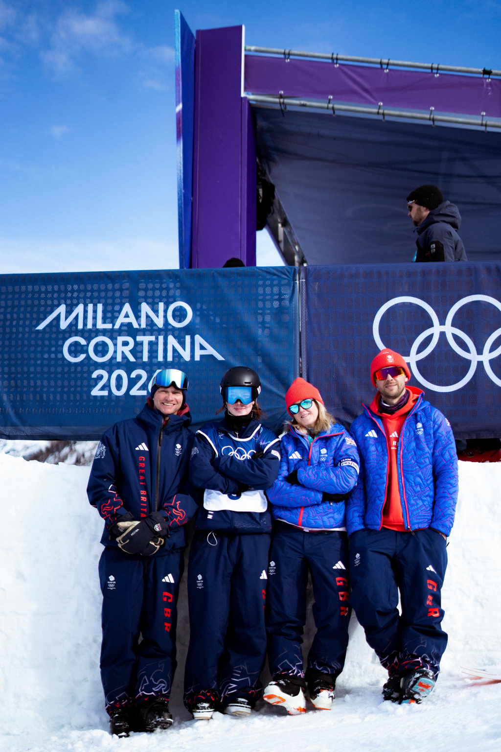 Four athletes in snow gear stand in front of a sign reading 'Milano Cortina 2026' with Olympic rings, snow and blue sky in the background at the Winter Olympics.