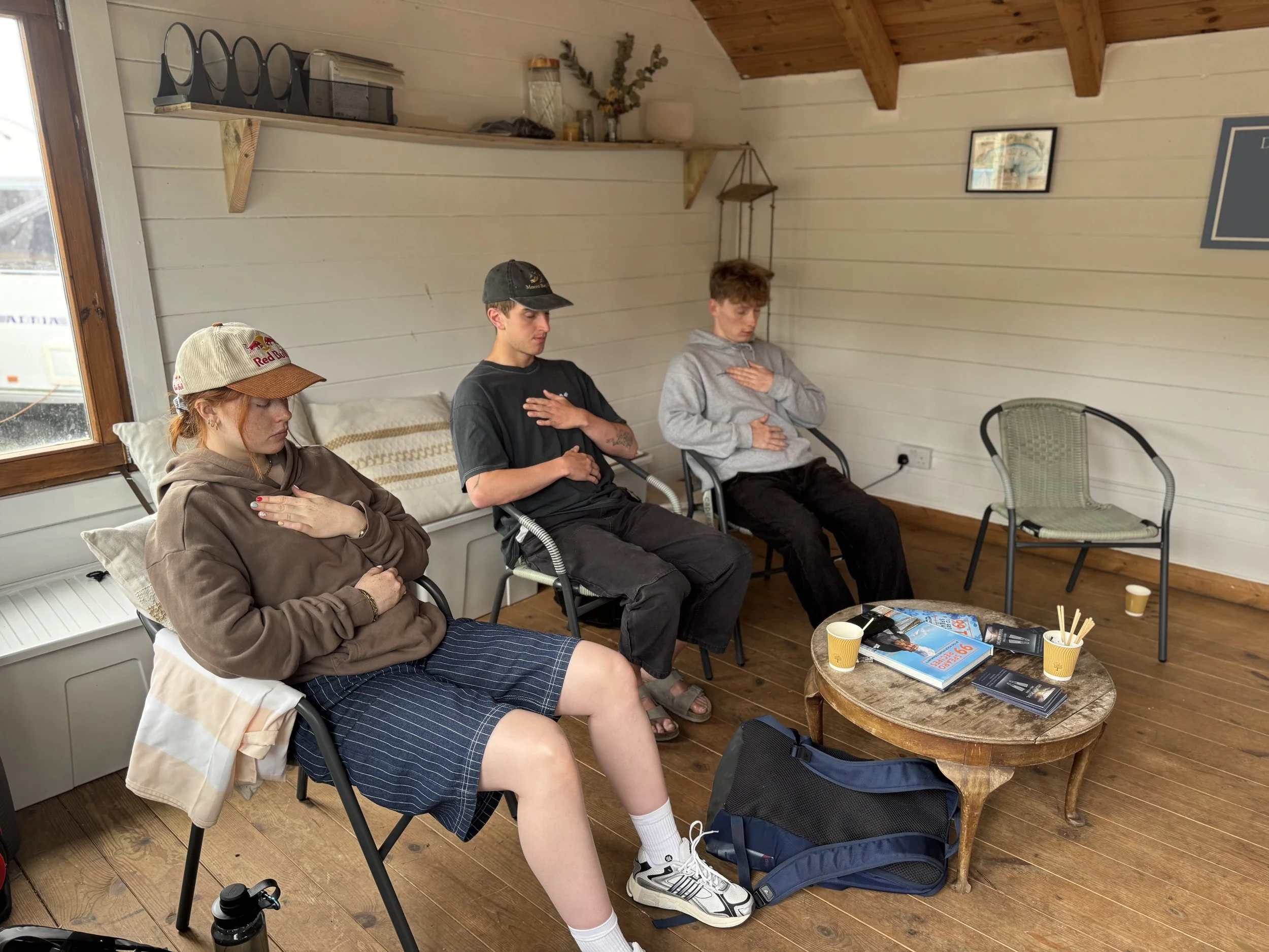 Three young adults seated in a room with wooden walls and ceiling, all with their hands on their chests and eyes closed, possibly meditating or participating in a mindfulness activity. A woman with red hair, wearing a red cap and brown hoodie, is seated in the foreground. Two young men, one with dark hair and a black cap, and the other with curly brown hair and gray hoodie, are seated beside her. A small wooden table with books, magazines, and cups is in front of them. Other chairs and a window with rain outside are visible.