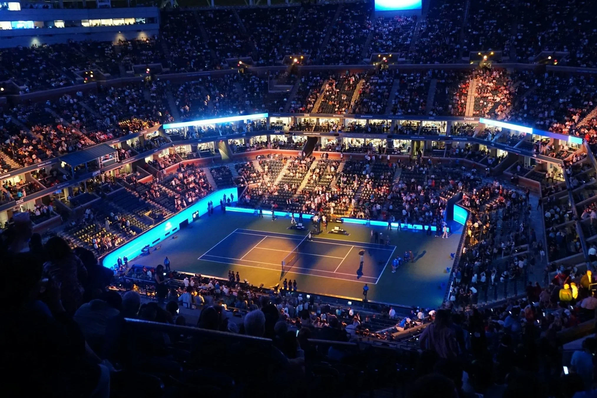 Indoor tennis stadium filled with spectators, illuminated courts, and a few players on the court preparing for a match.