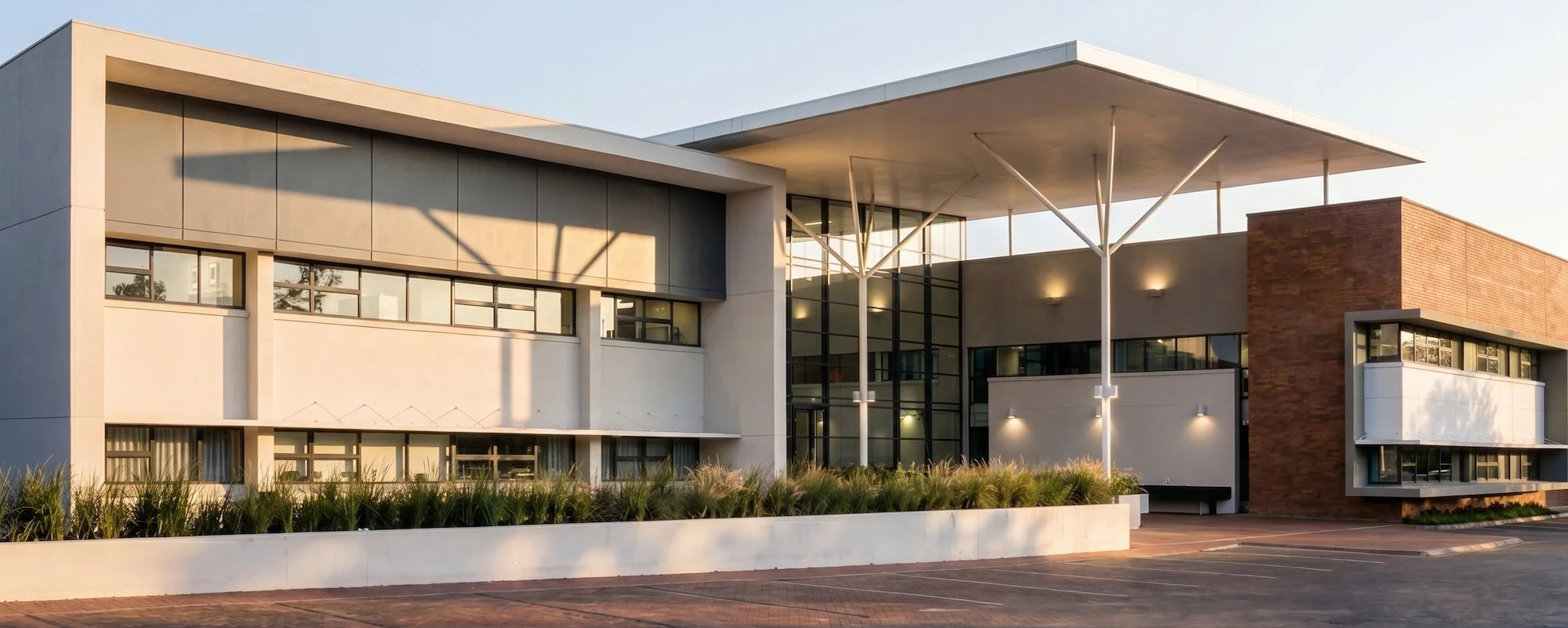 Modern commercial building with large glass windows, white and brick exterior, and a prominent flat canopy supported by thin slanted columns, set against a clear sky.