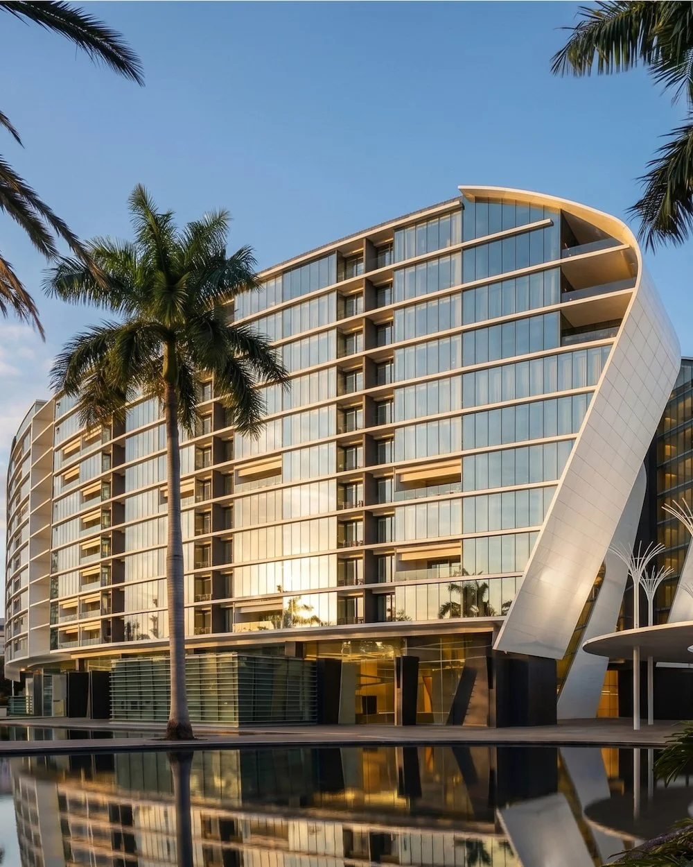 Modern glass hotel building with palm trees and a water reflection in a tropical setting.