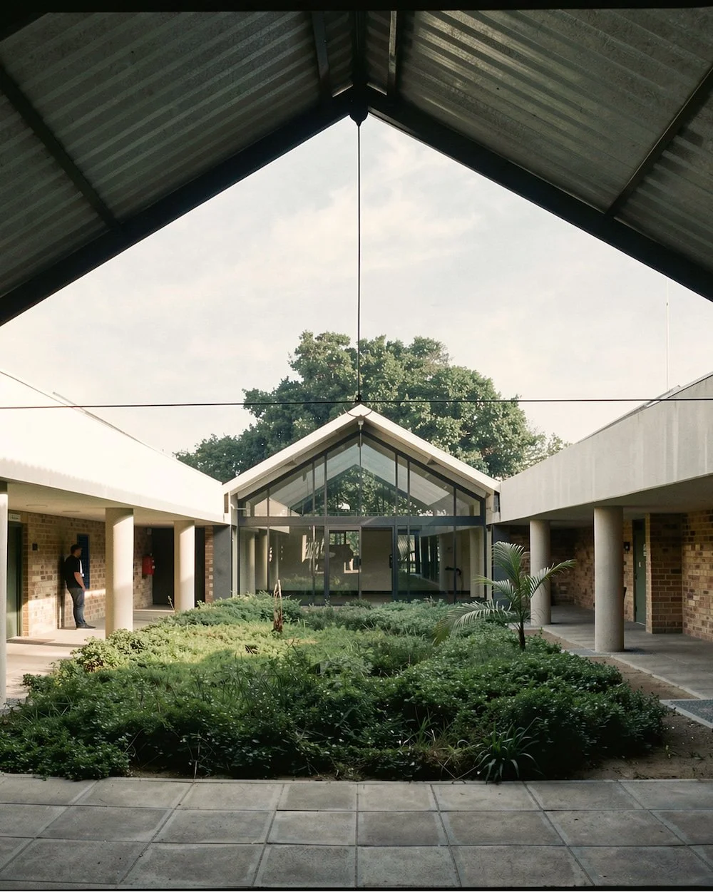 View of a courtyard with garden beds surrounded by a building with modern architecture, brick walls, white concrete supports, and large glass windows; the roof is pitched with a triangular peak.