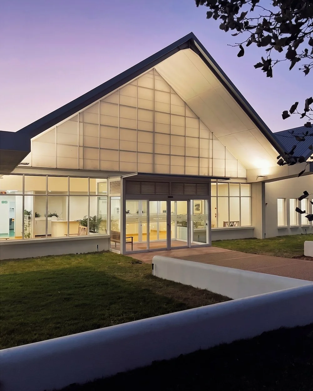 Modern building with large glass windows and a triangular roof, illuminated outside during sunset, surrounded by a small grassy area.
