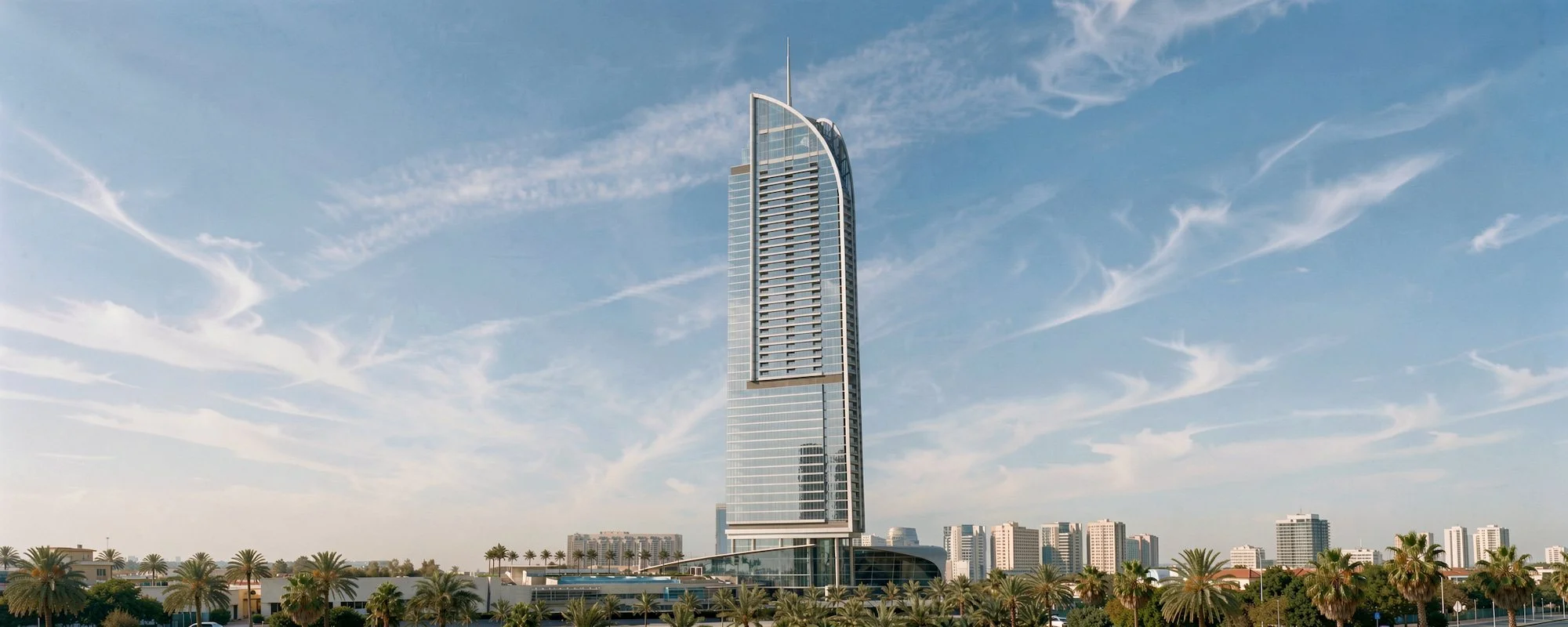 A tall, modern skyscraper with a curved top, situated in a city with palm trees and shorter buildings at its base, under a blue sky with wispy clouds.
