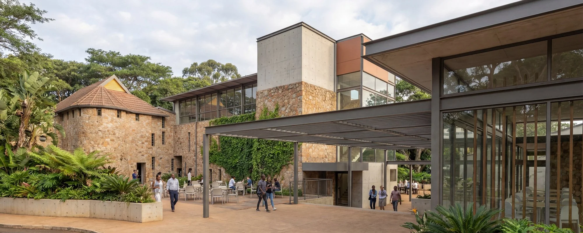 Modern building with glass walls, stone and concrete exterior, surrounded by greenery, with people walking and sitting outside.