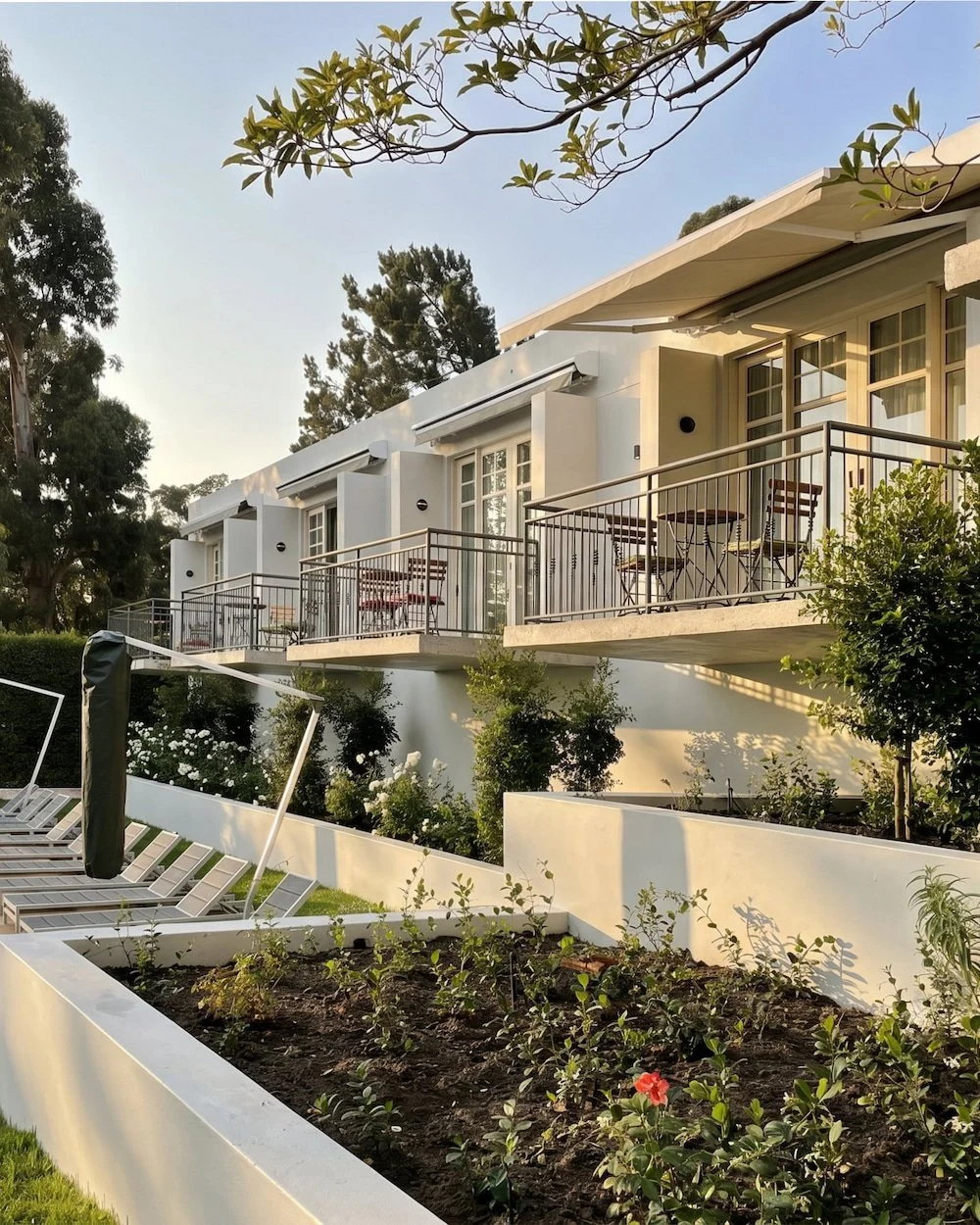 Modern white apartment building with balconies, outdoor seating, and a garden area with plants and flowers in the foreground, during late afternoon sunlight.