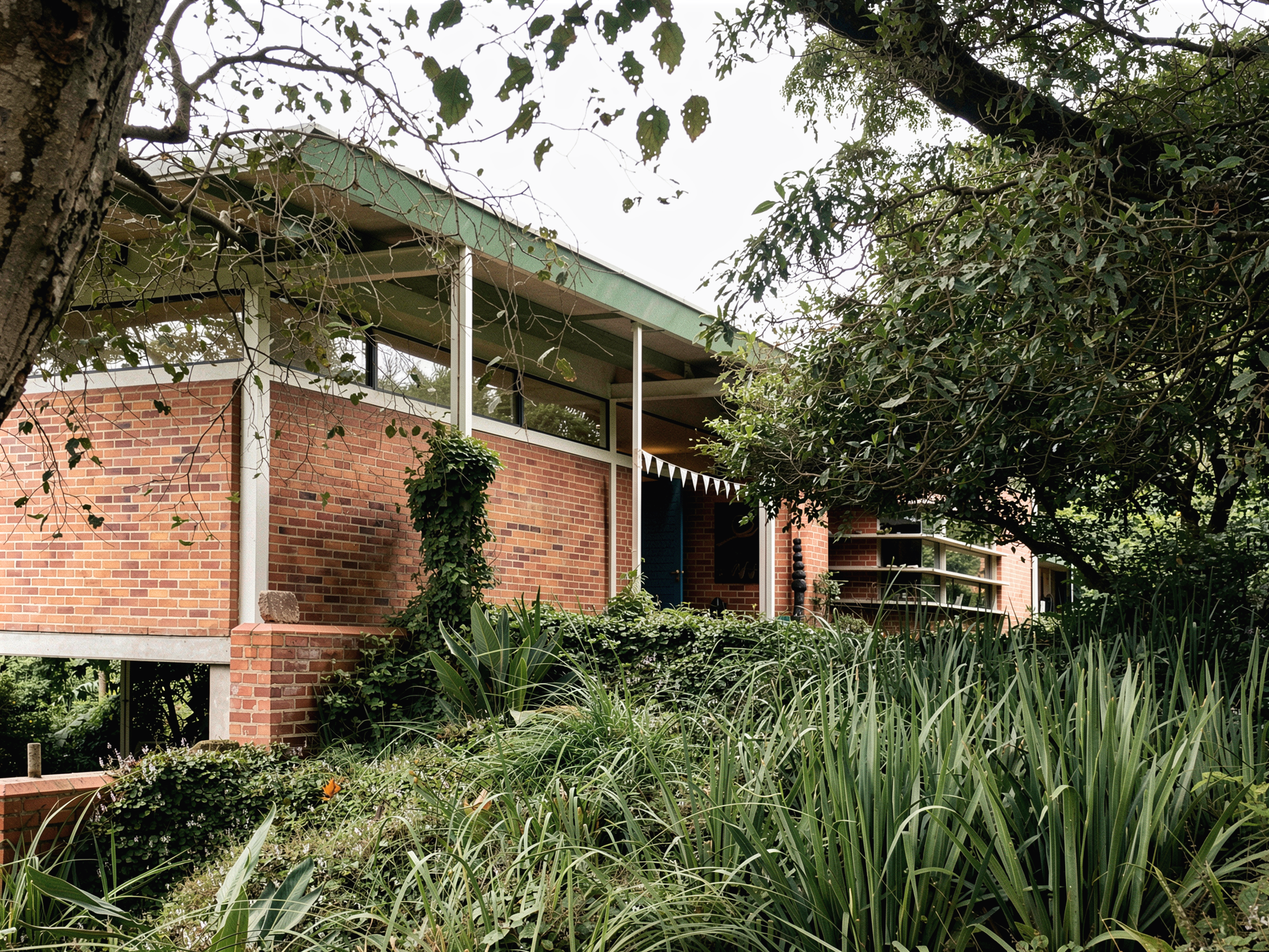 A modern house with a brick exterior, large windows, and a green roof, surrounded by lush greenery and trees.