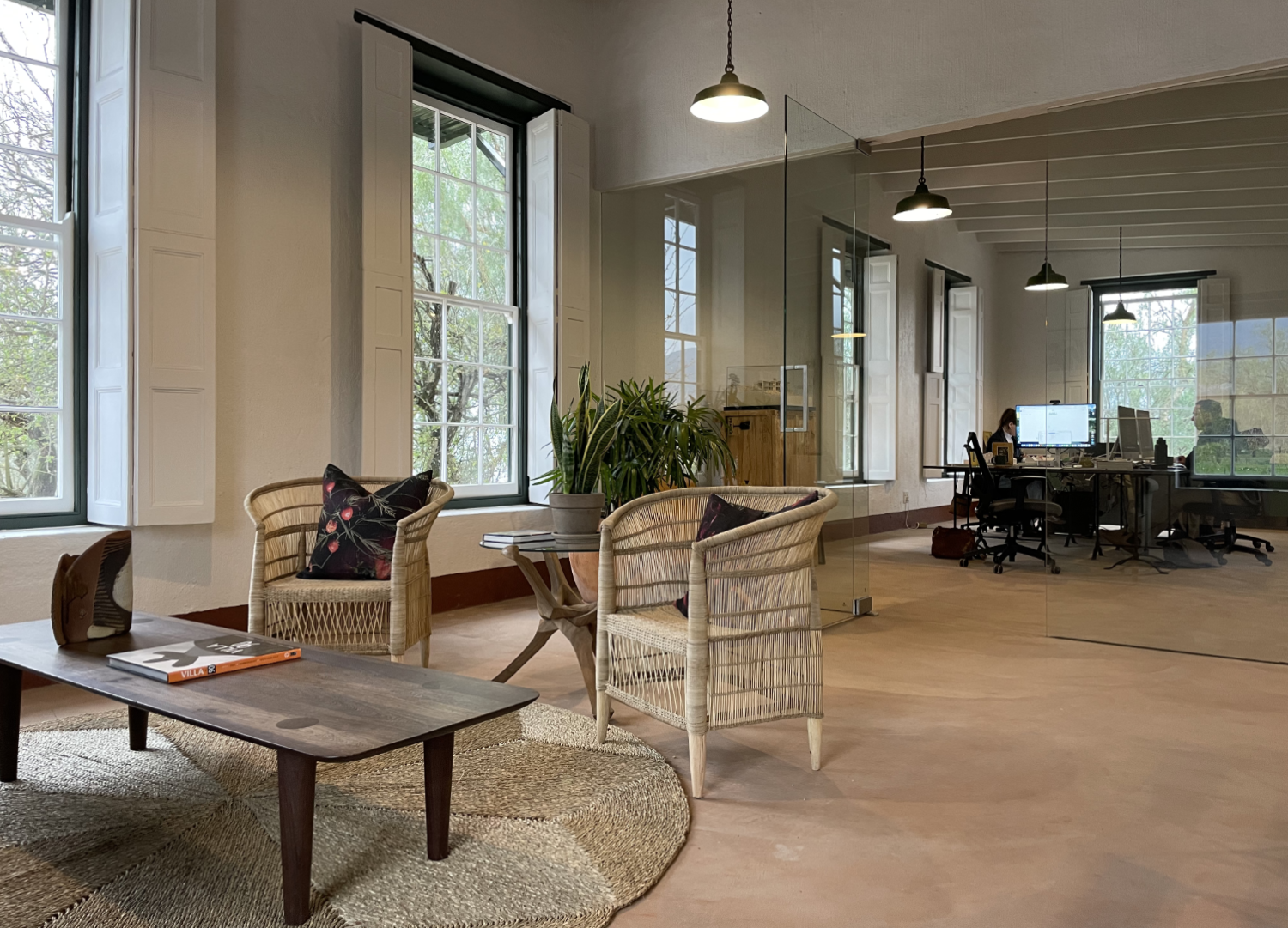 Office space with large windows, wicker chairs with cushions, a plant on a side table, a wooden coffee table, and a glass wall separating the sitting area from workstations with computers and people working.