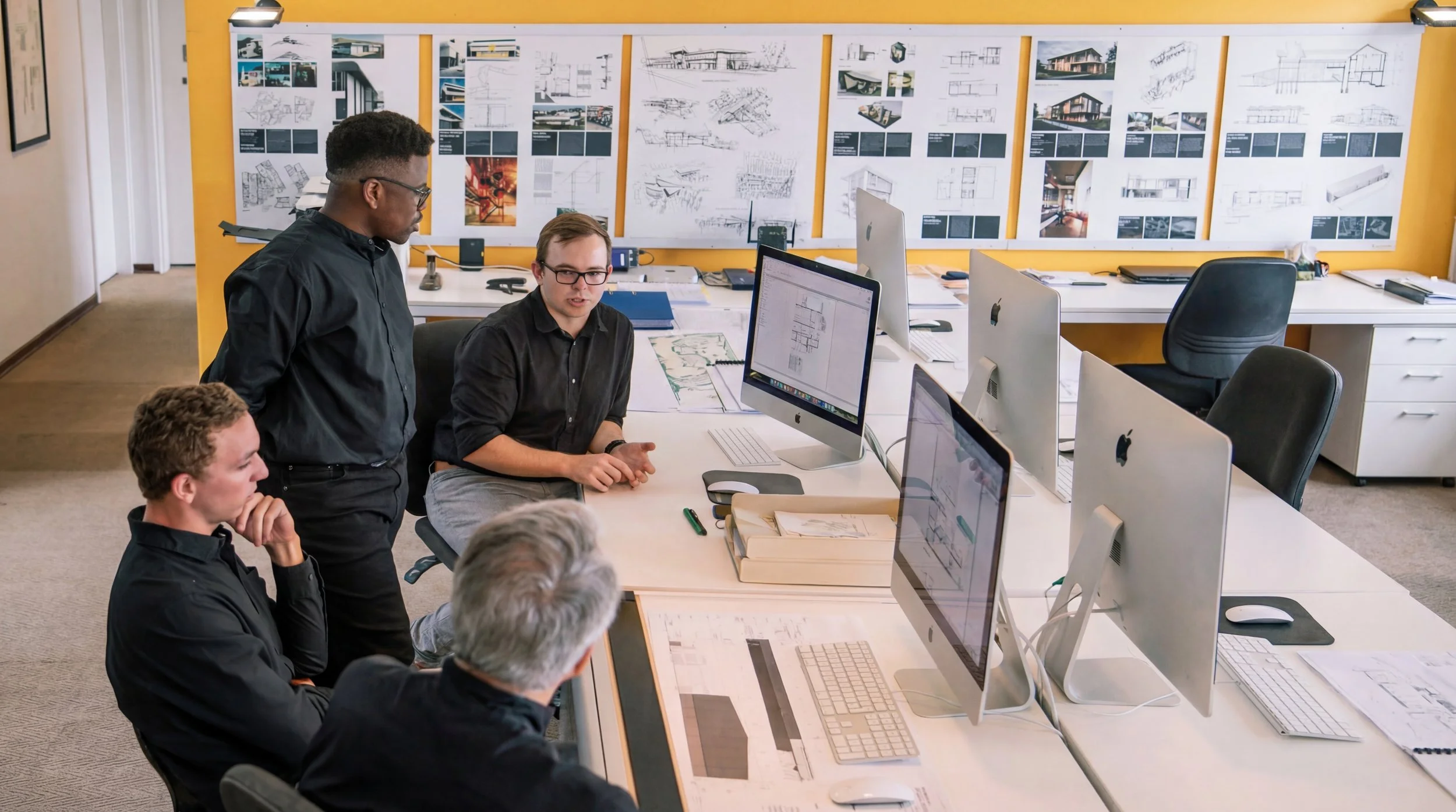 A group of four men working in an architecture office with blueprints and CAD designs on their desktop computers. A yellow wall behind them features a large board displaying architectural plans and drawings.