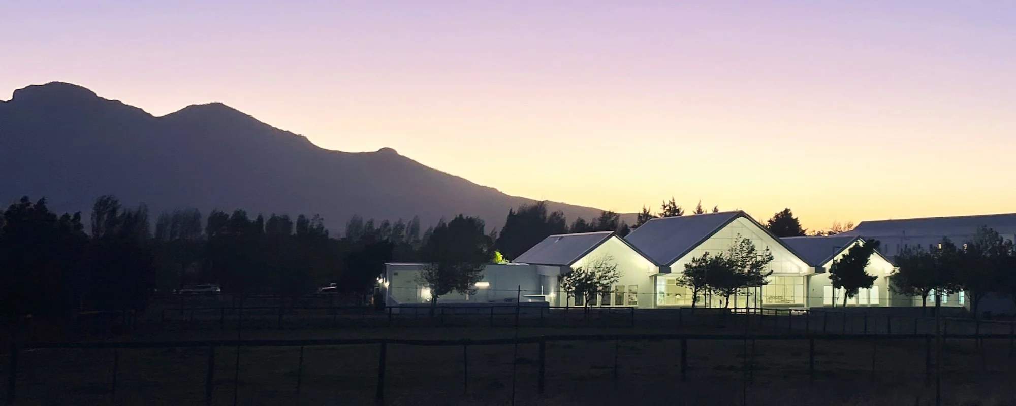 Modern building with glass walls illuminated at dusk, set against a mountain range with a colorful sky