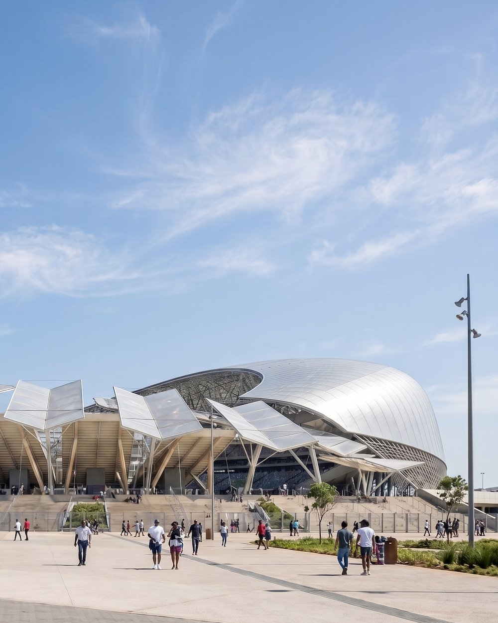 Modern architectural building with curved silver exterior and white solar panel-like structures in front, pedestrians walking outside on a sunny day with a blue sky and white clouds.