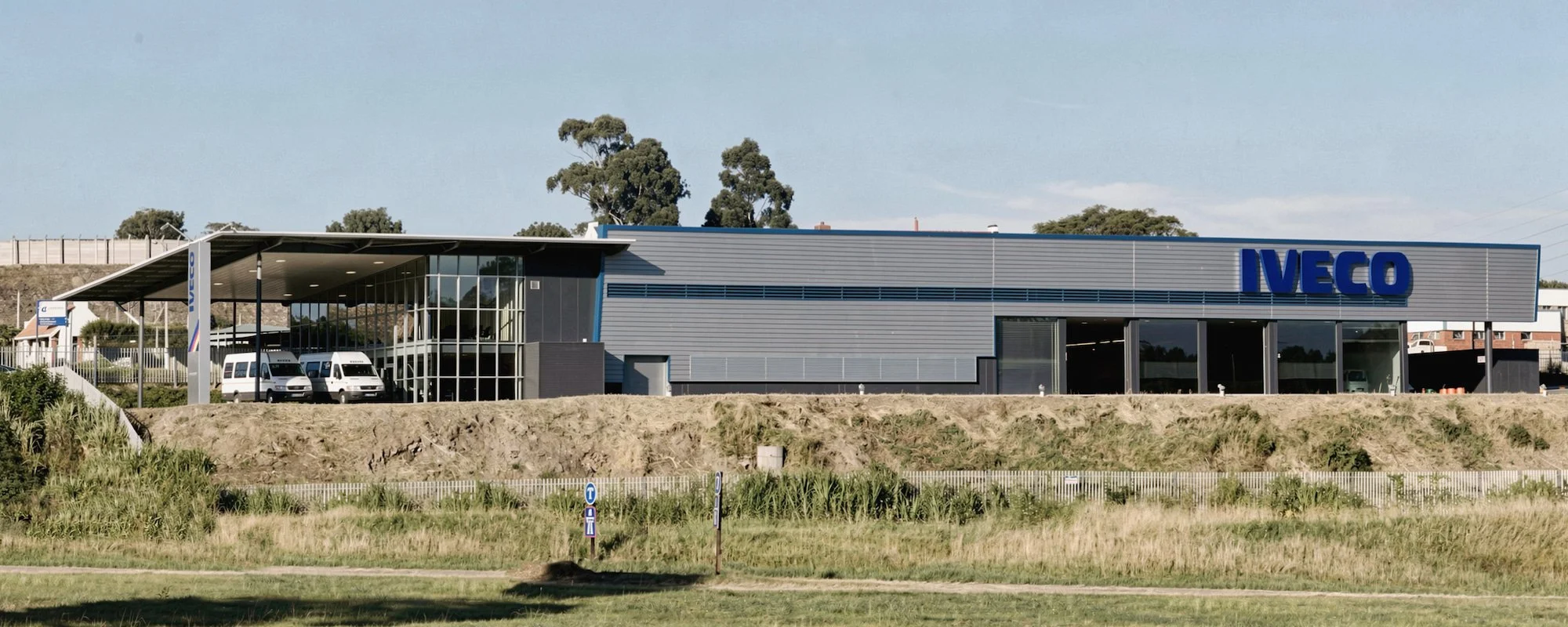 Exterior of an Iveco dealership or service center with a modern design, multiple white vans parked under a large canopy, and the Iveco logo on the building's facade.