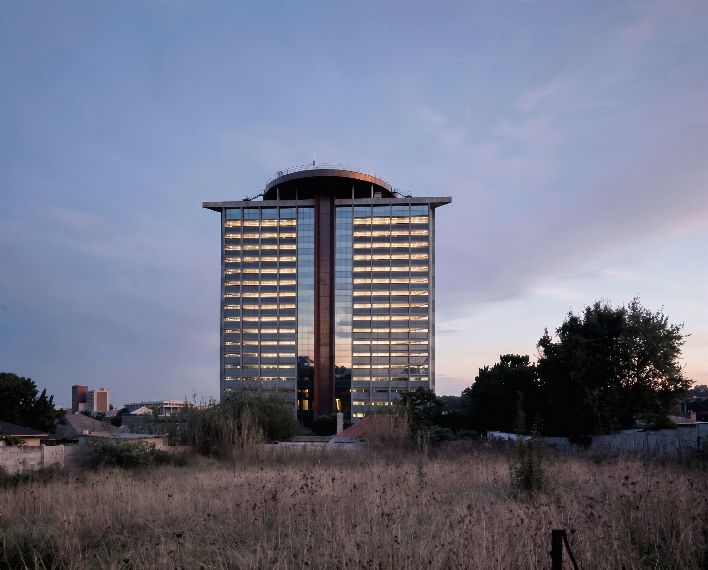 A tall modern building with glass windows and a rounded top, surrounded by trees and smaller buildings, under a cloudy sky during dusk.