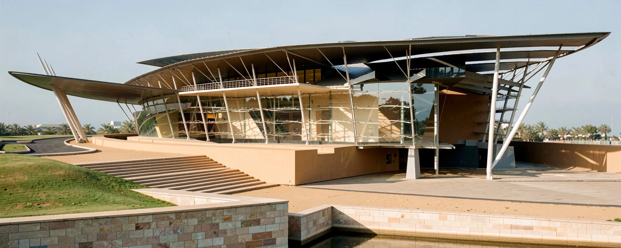 Modern architectural building with large glass windows, multiple overhanging roofs, and white support beams, set against a background of palm trees and a clear sky.