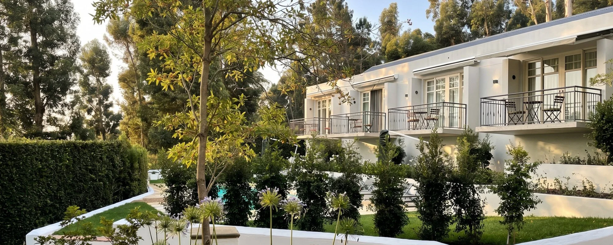 Modern white apartment building with balconies surrounded by lush green trees and plants, with some outdoor furniture visible on the balconies.