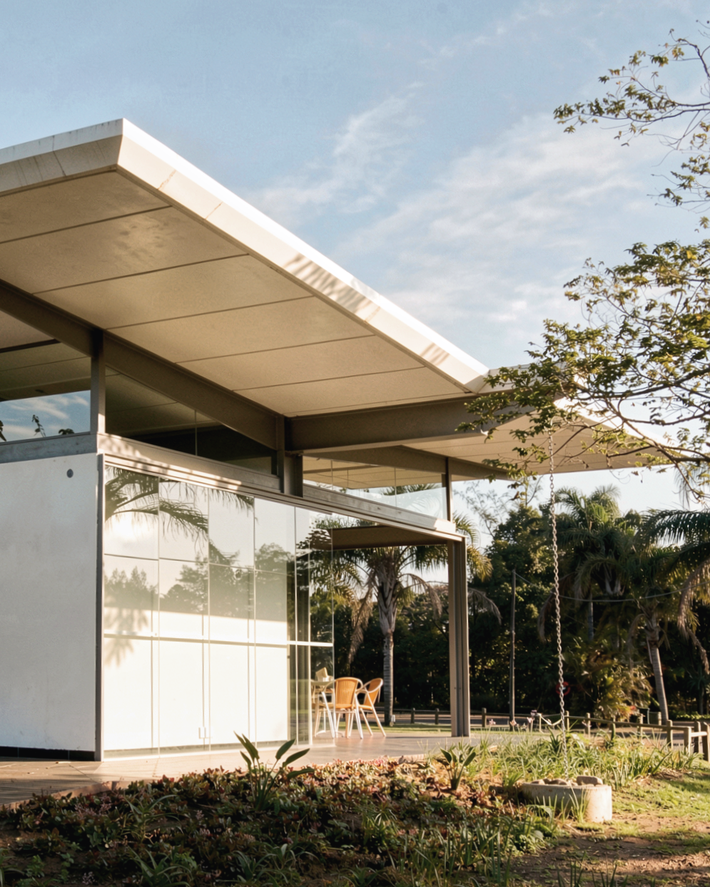 Modern house with large glass windows and a sloped roof, surrounded by greenery and palm trees, with a swing hanging from a tree and outdoor table and chairs.