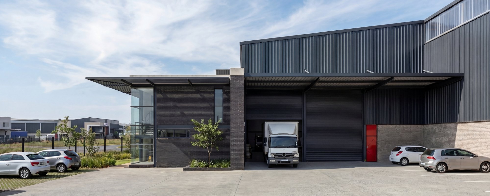 Modern industrial warehouse building with black metal siding, large garage door, and a red entry door. Several parked cars and a delivery truck are in front, with a landscaped area and trees, under a partly cloudy sky.