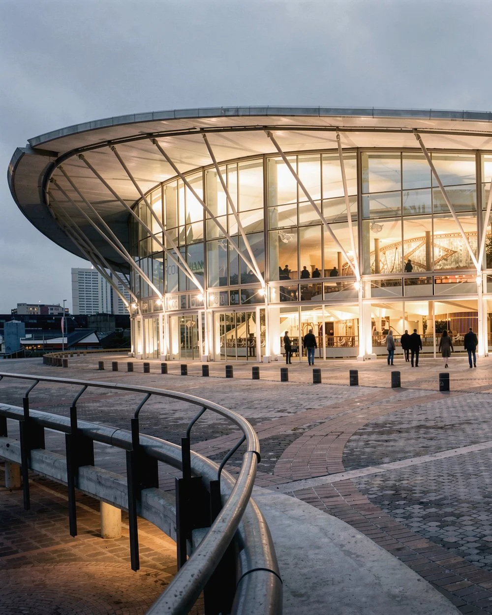 Modern glass building with interior lighting and people inside, evening setting, curved architecture, cityscape background.