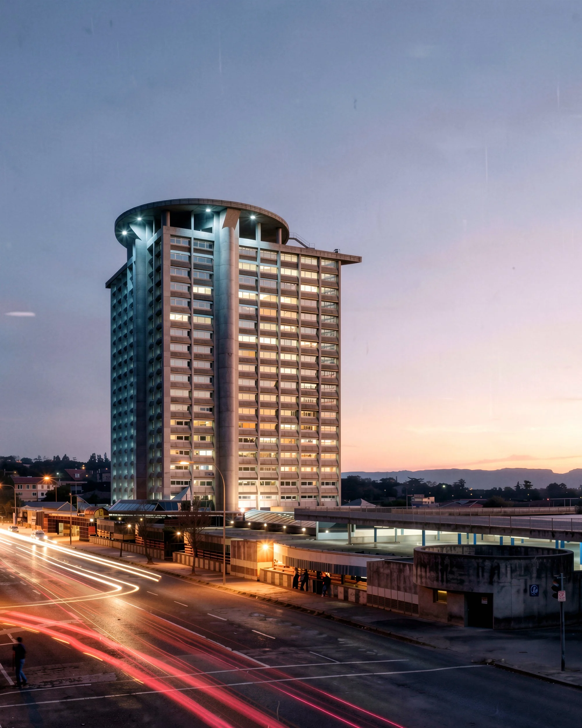 Cityscape at dusk featuring a tall modern office building with illuminated windows, a peaceful sky with subtle hues of sunset, and light trails from moving vehicles on the street below.