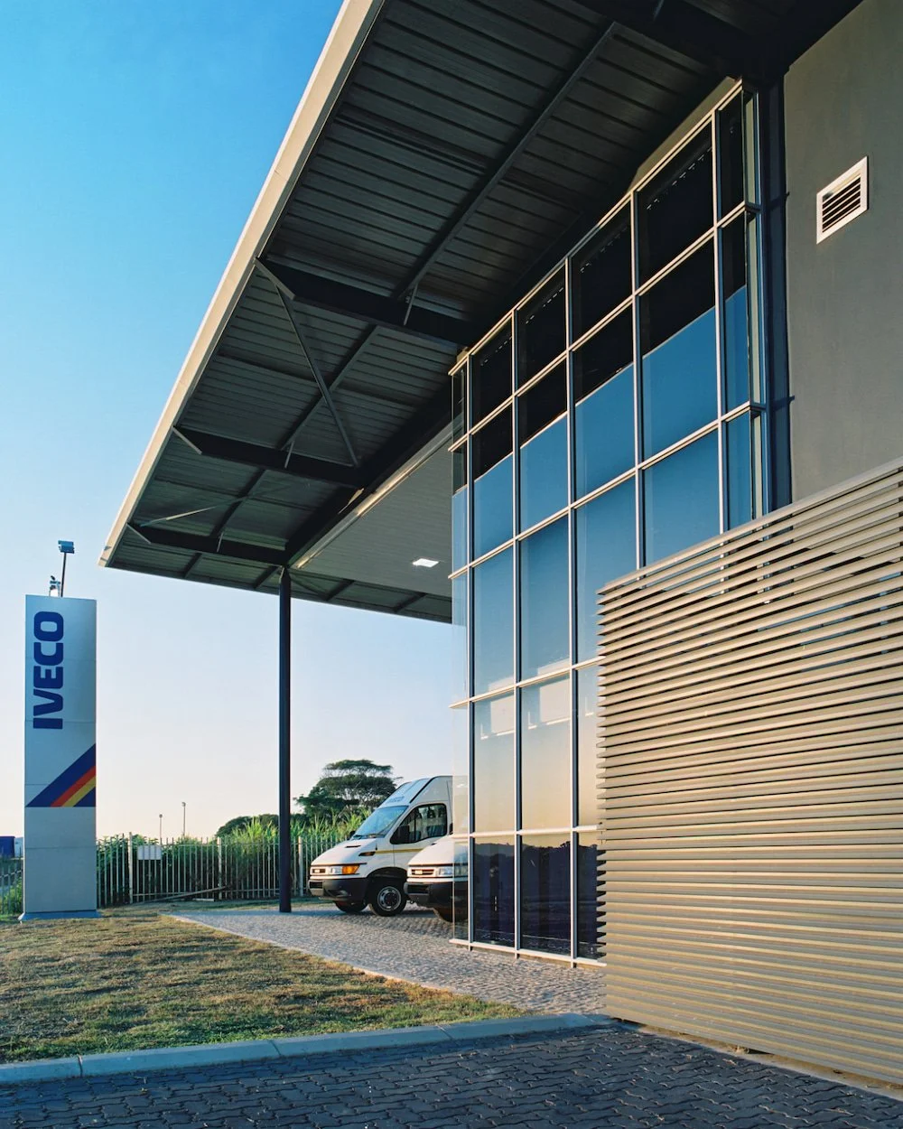 Modern building with glass windows, a large overhanging roof, and two parked white vans outside. IVECO sign visible near the building.