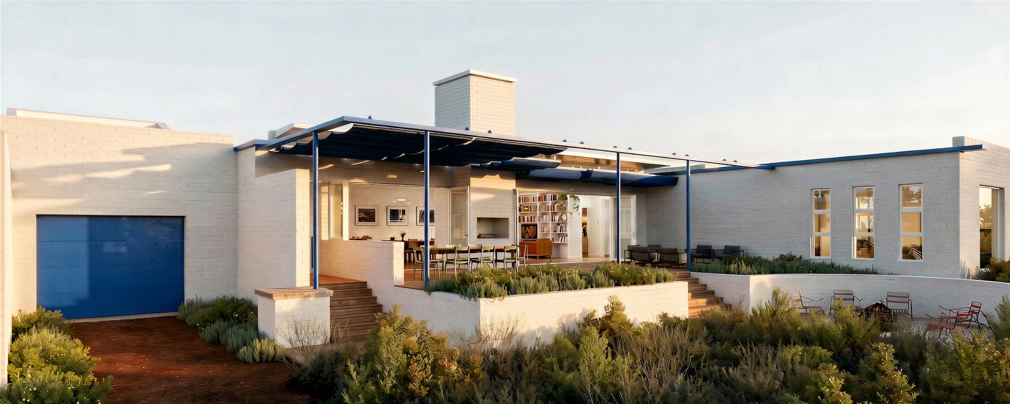 Modern house with white brick exterior, blue accents, and outdoor seating area with greenery, on a sunny day.