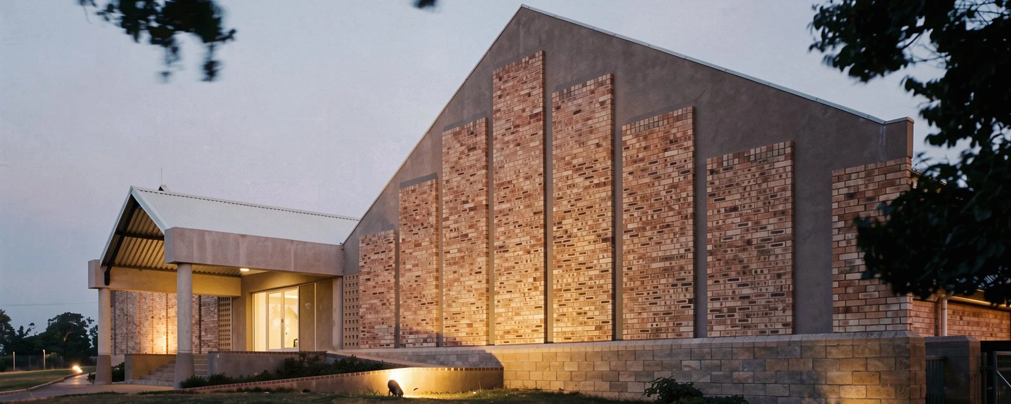 A modern building with brick accents and a sloped roof, illuminated at dusk, with trees nearby.