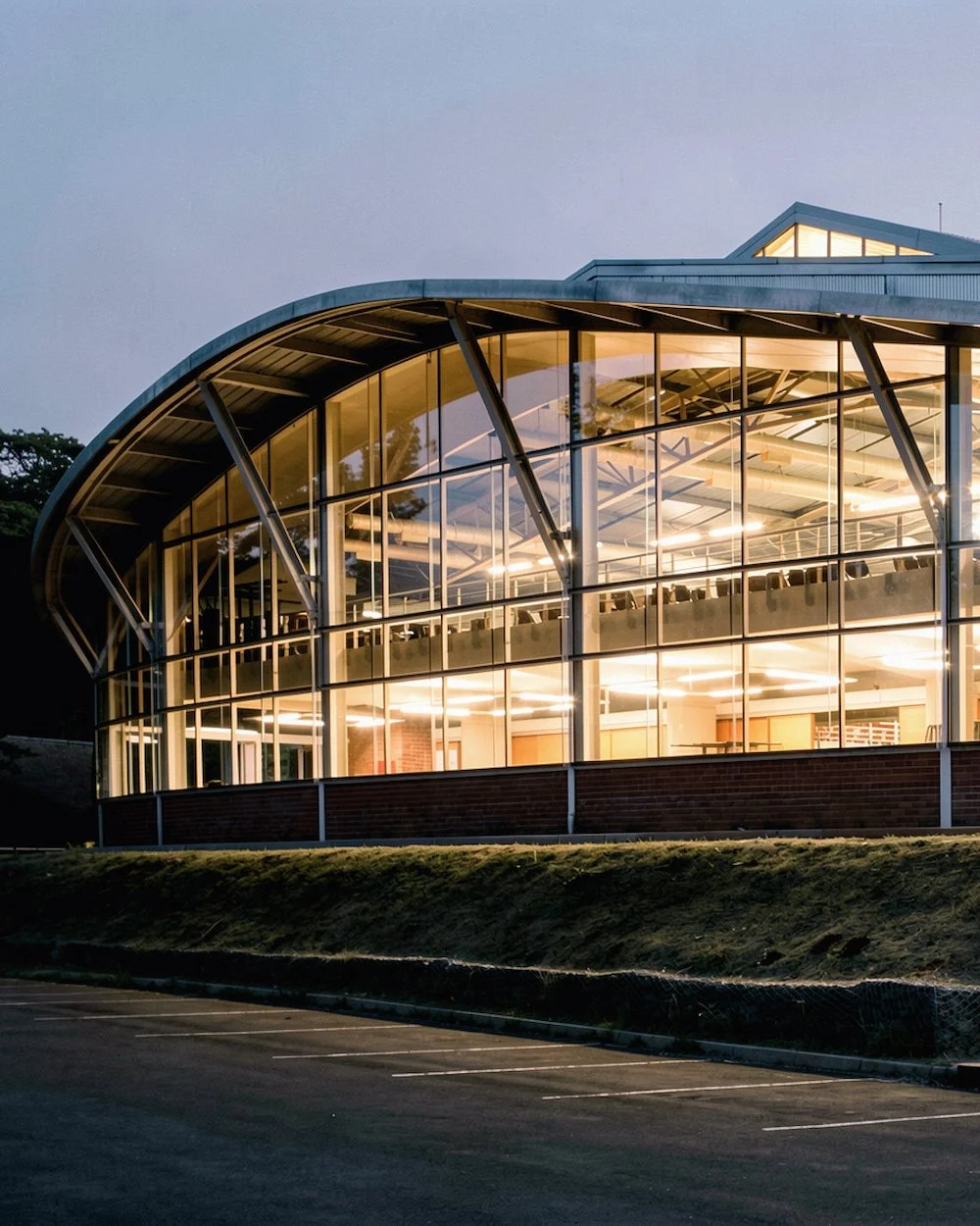 Modern glass building with illuminated interior, curved roof, and large windows, situated next to an empty parking lot at dusk.