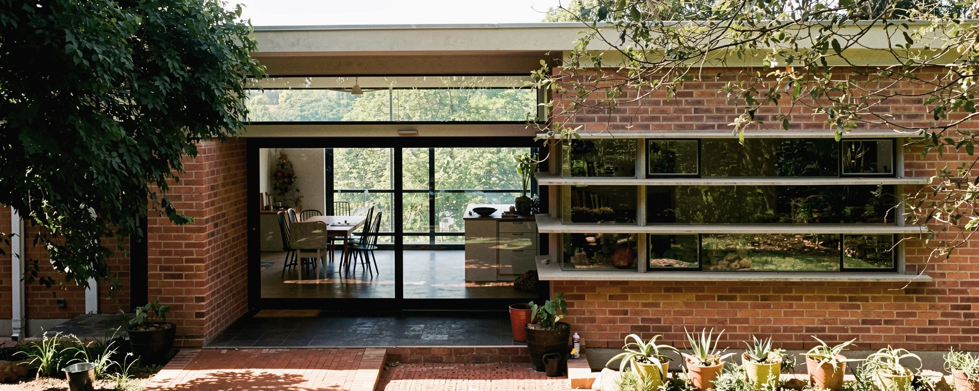 View of a modern house with a brick exterior, large sliding glass doors, and an indoor dining area with a wooden table and chairs visible through the glass. There are potted plants and trees outside, with sunlight creating shadows on the brick patio.