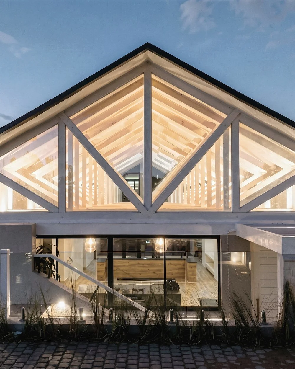 A modern house with a glass front and a triangular, illuminated wooden gabled roof structure at dusk. The house features large windows and warm interior lighting.