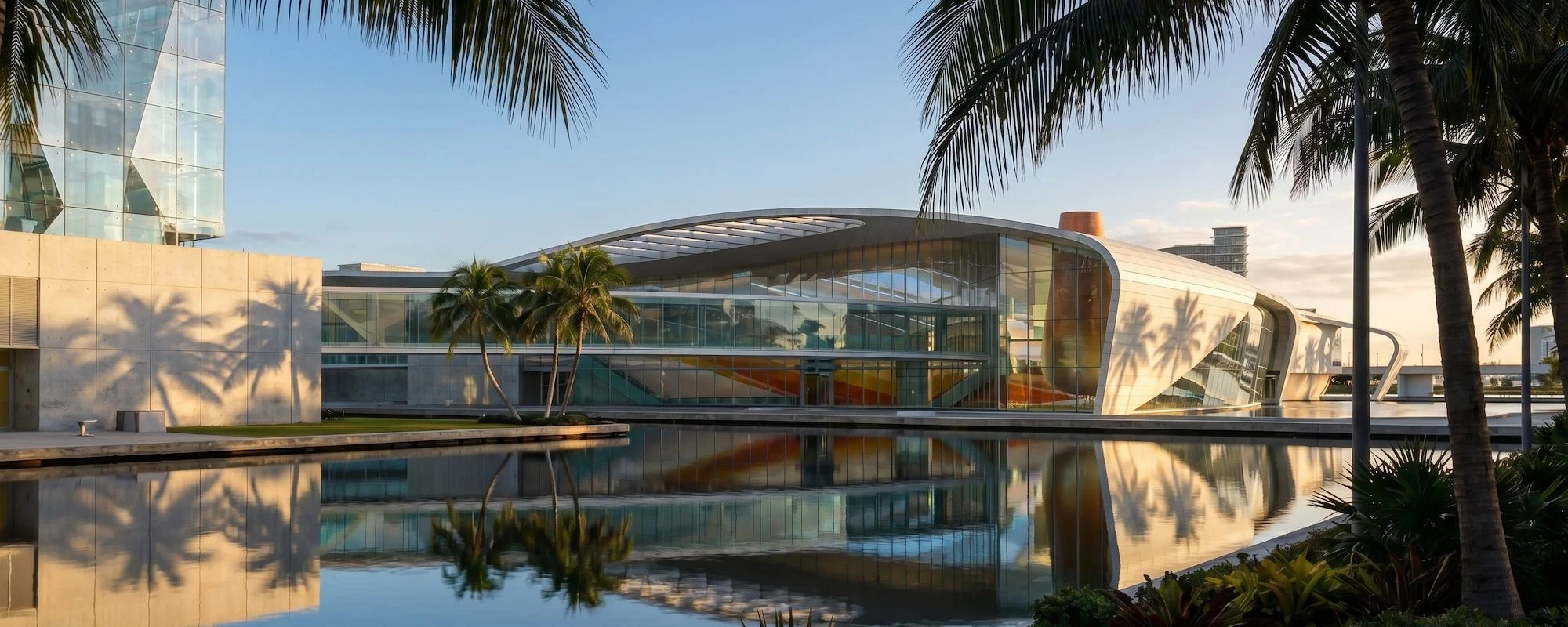 Modern building with curved glass architecture, surrounded by palm trees and a water reflection, during sunset.