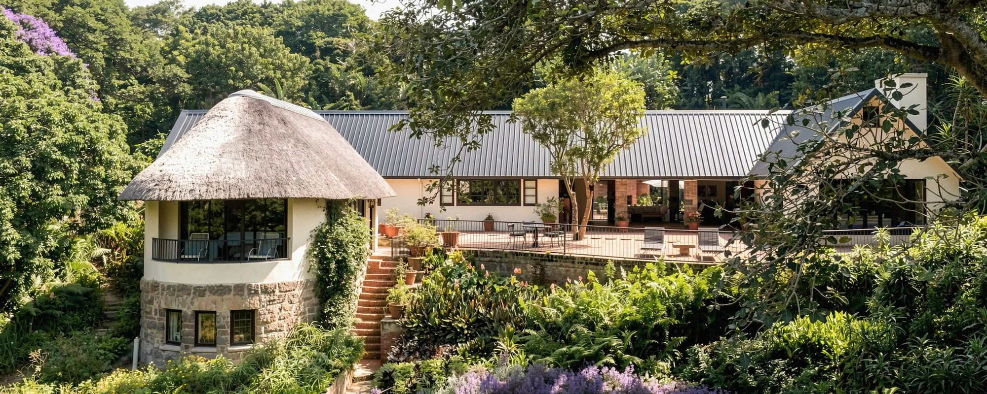 A large house with a metal roof surrounded by lush greenery and trees, featuring a spacious outdoor deck with patio furniture and potted plants, with vibrant flowering bushes in the foreground.