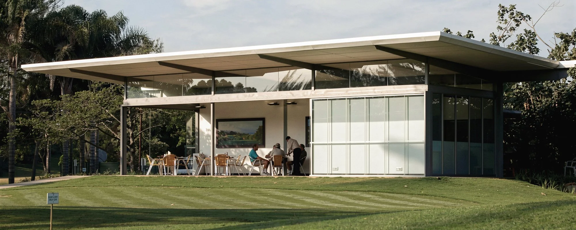 Modern building with glass walls, outdoor seating, and a group of people inside, surrounded by trees and a well-maintained lawn.