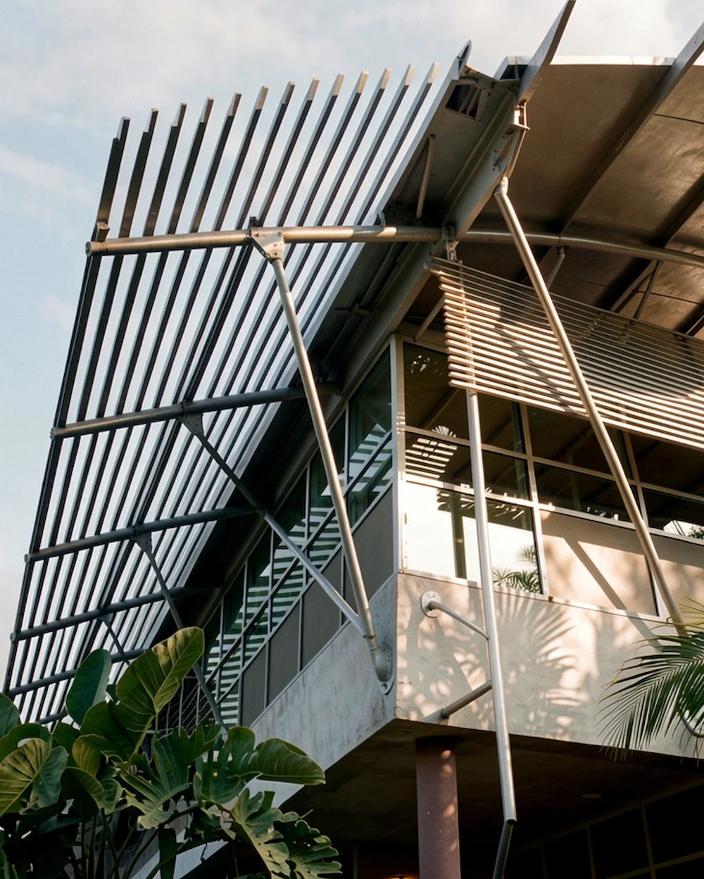 Close-up of a modern building with metal sunshade panels, glass windows, and metal support beams, with tropical plants in the foreground.