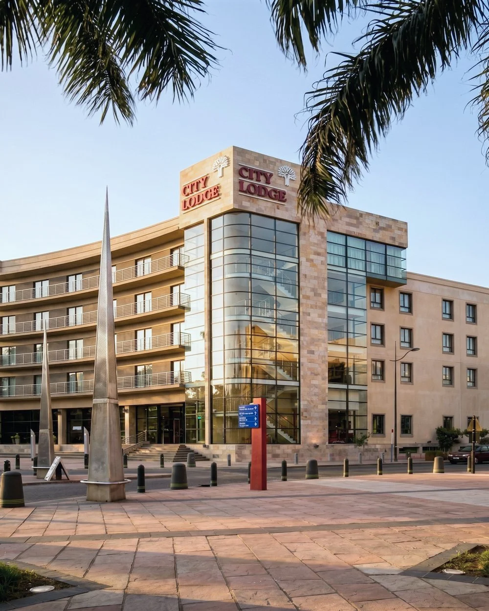 Modern Hilton City Lodge building with glass facade, palm trees, and stone accents, located on a city street with sidewalks and bollards.